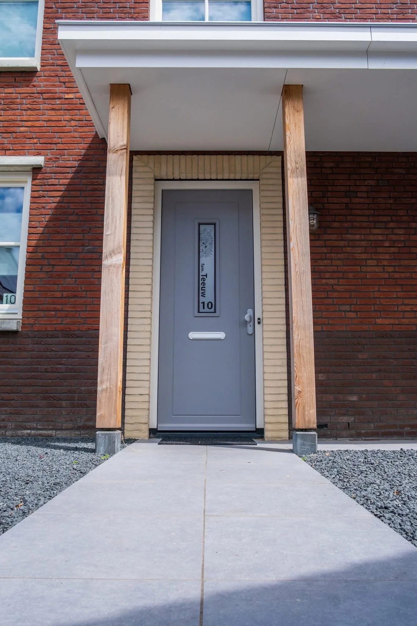 Front door of a modern brick apartment building with a gray door, small window, and a vertical sign with the apartment number '10' and the name 'Teeuw' on it. The entrance is framed by two unfinished wooden posts and a small porch with concrete floor