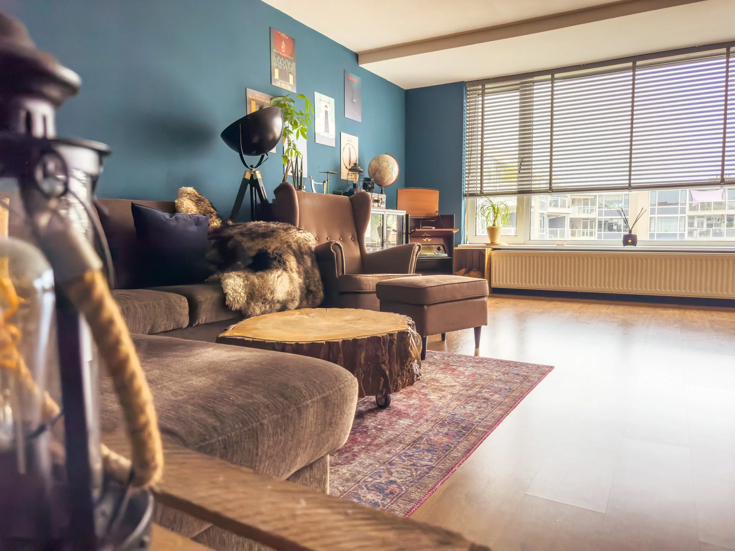 Living room with large window, blue wall, brown couch with pillows and fur throw, wooden coffee table, armchair, potted plants, vintage globe, framed art on wall, radiator under window, patterned rug, wooden floor.