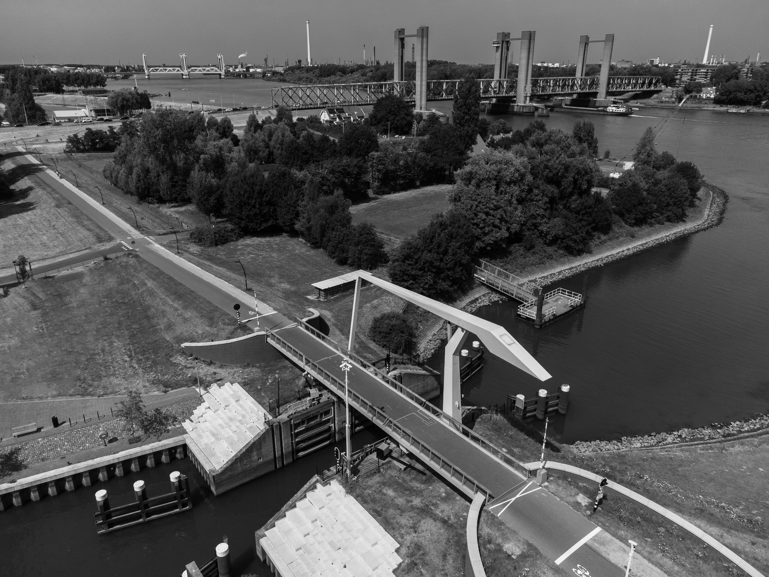 Black and white aerial photo of a park with a drawbridge over a river, surrounded by trees and industrial buildings in the background.