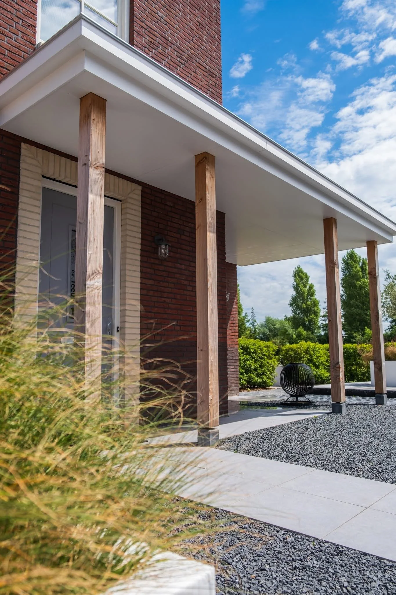 Modern house with a covered porch supported by wooden posts, brick exterior walls, decorative outdoor lighting, lush green landscaping, and a clear blue sky with some clouds.