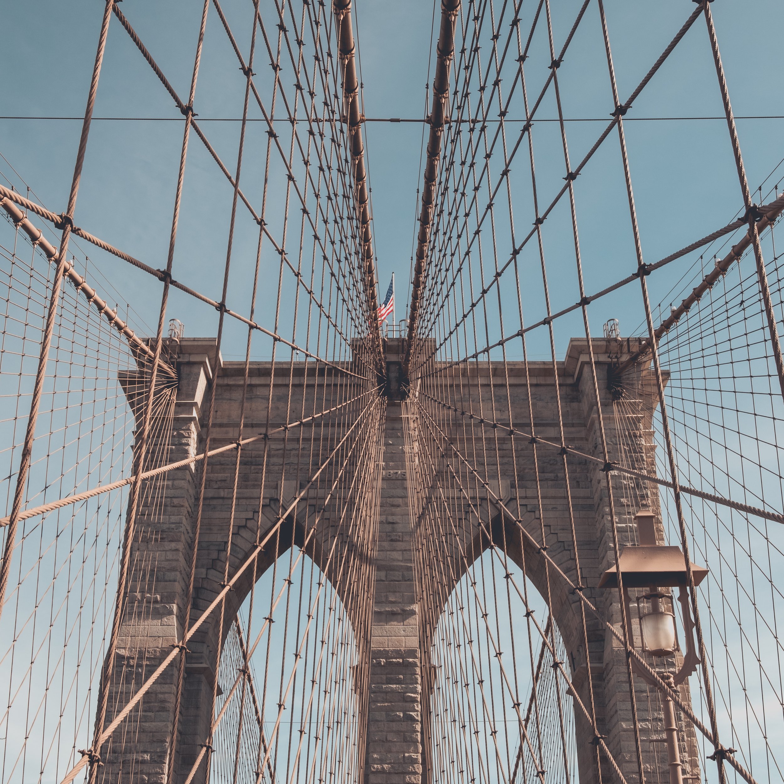 View from below of the Brooklyn Bridge with its stone towers, steel cables, and American flag against a blue sky.