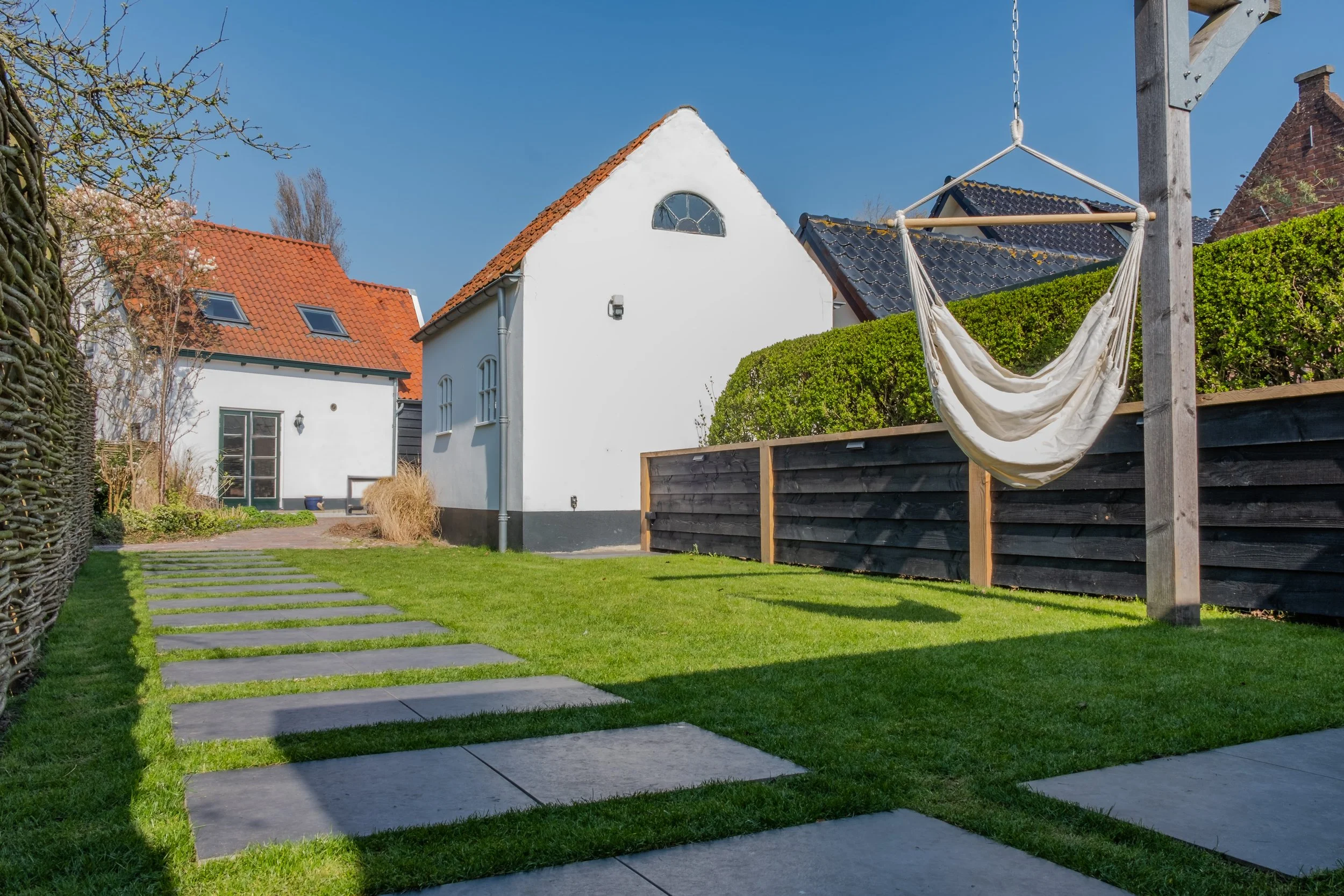 A backyard with a paved stepping stone pathway on a lush green lawn, a white house with a red tiled roof, a black fence, a wooden post with a hanging hammock, and neighboring houses under a clear blue sky.