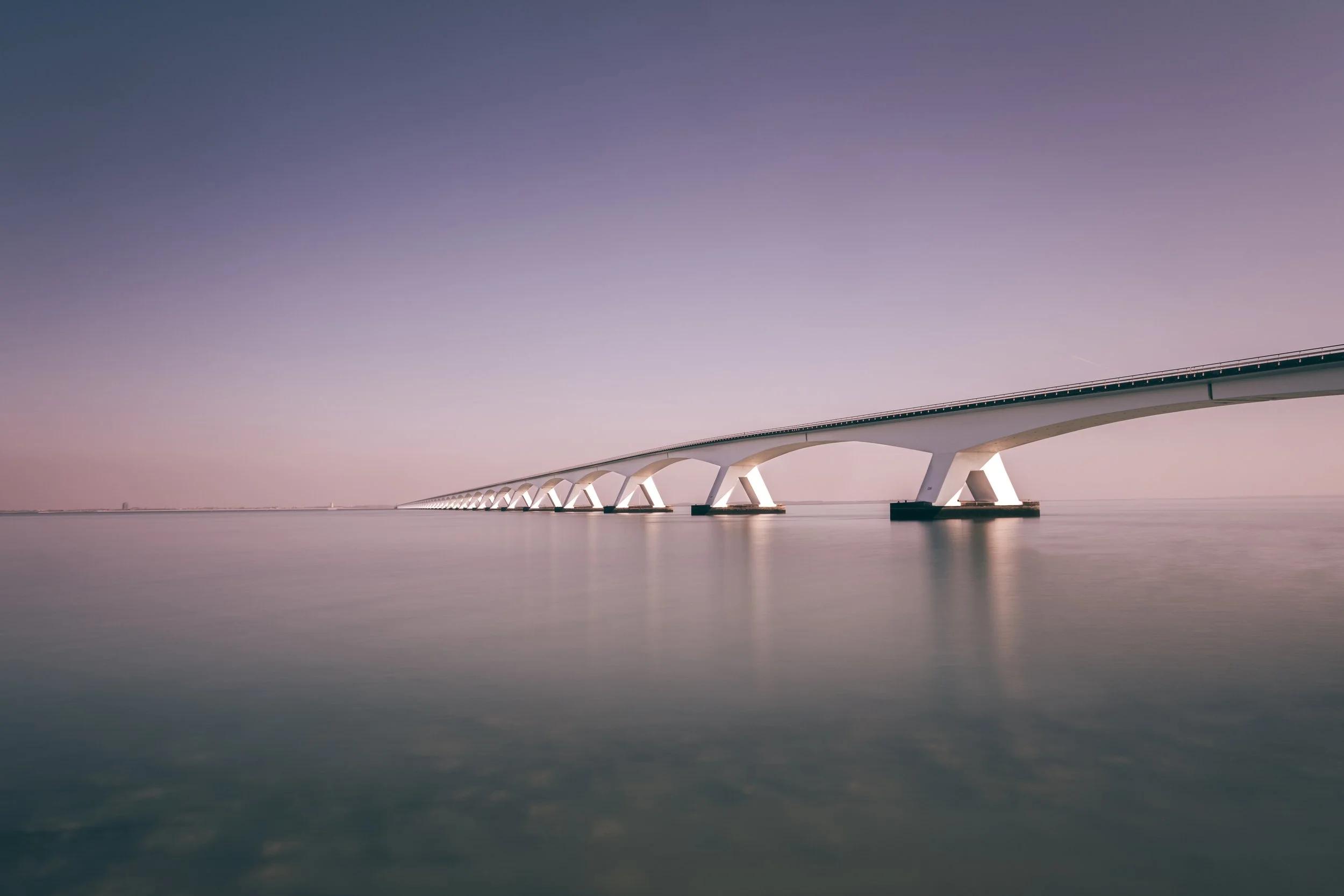 A long white bridge extending over calm water under a gradient sky with purple and pink hues.