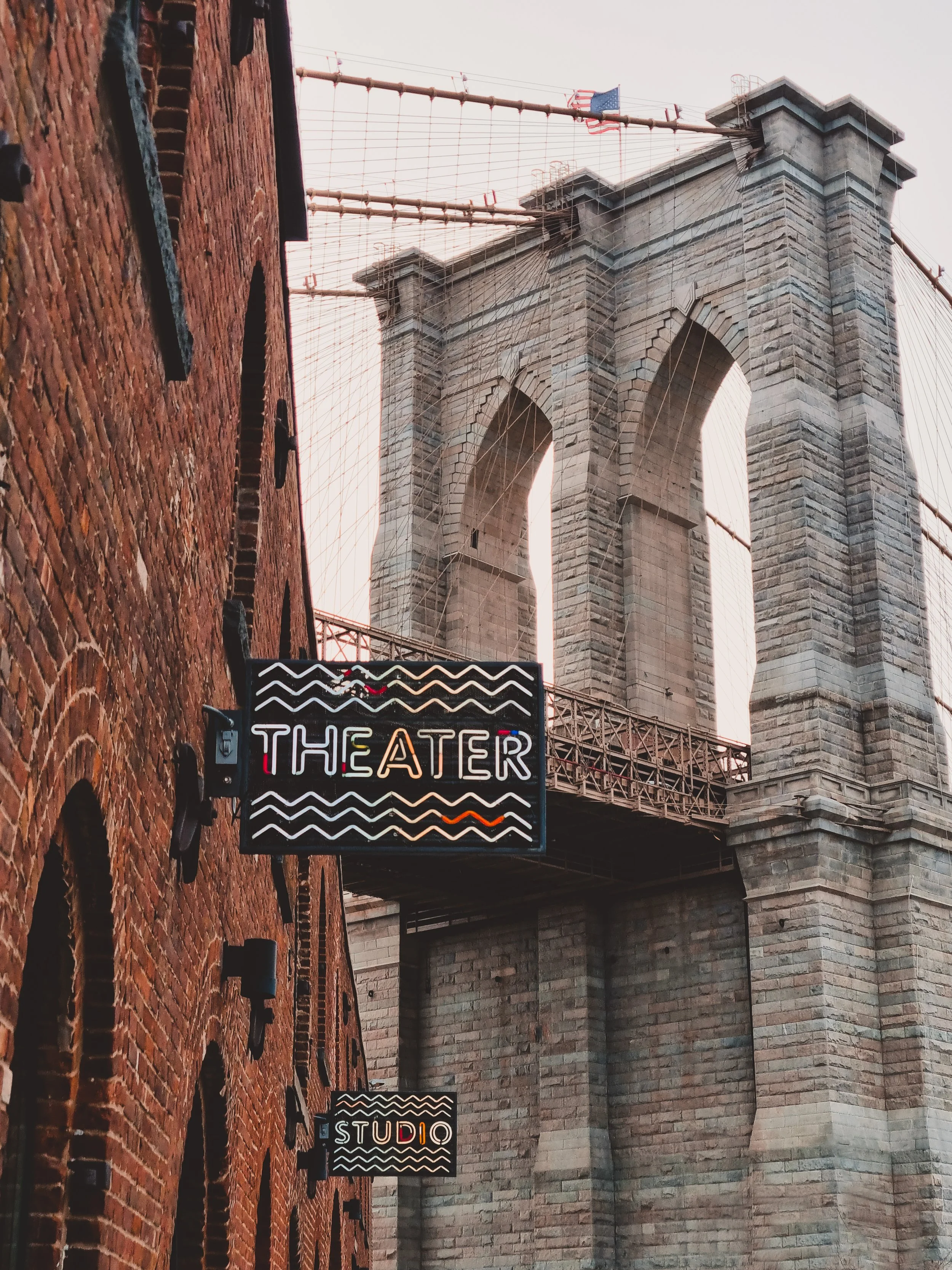Neon signs displaying 'THEATER' and 'STUDIO' in front of the Brooklyn Bridge with its iconic stone towers and suspension cables in New York City.
