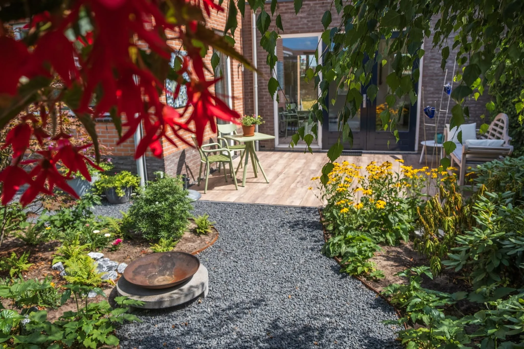 A cozy backyard garden with a gravel pathway and lush green plants. There is a wooden deck with patio furniture, including a small table with chairs, a bench, and decorative planters. The scene is shaded by overhanging leaves from trees or bushes.
