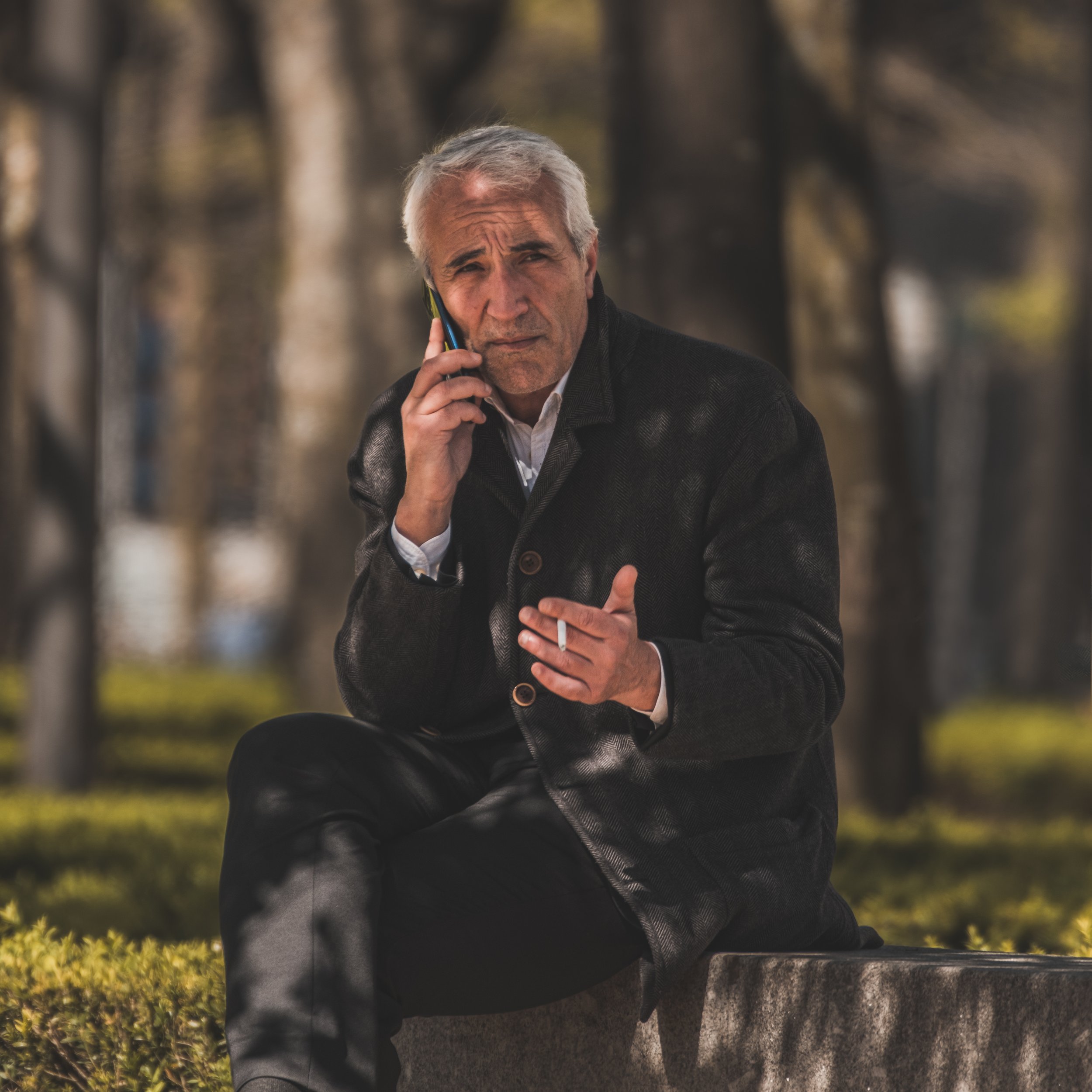 An elderly man with gray hair sitting on a stone bench outdoors, talking on his cellphone.