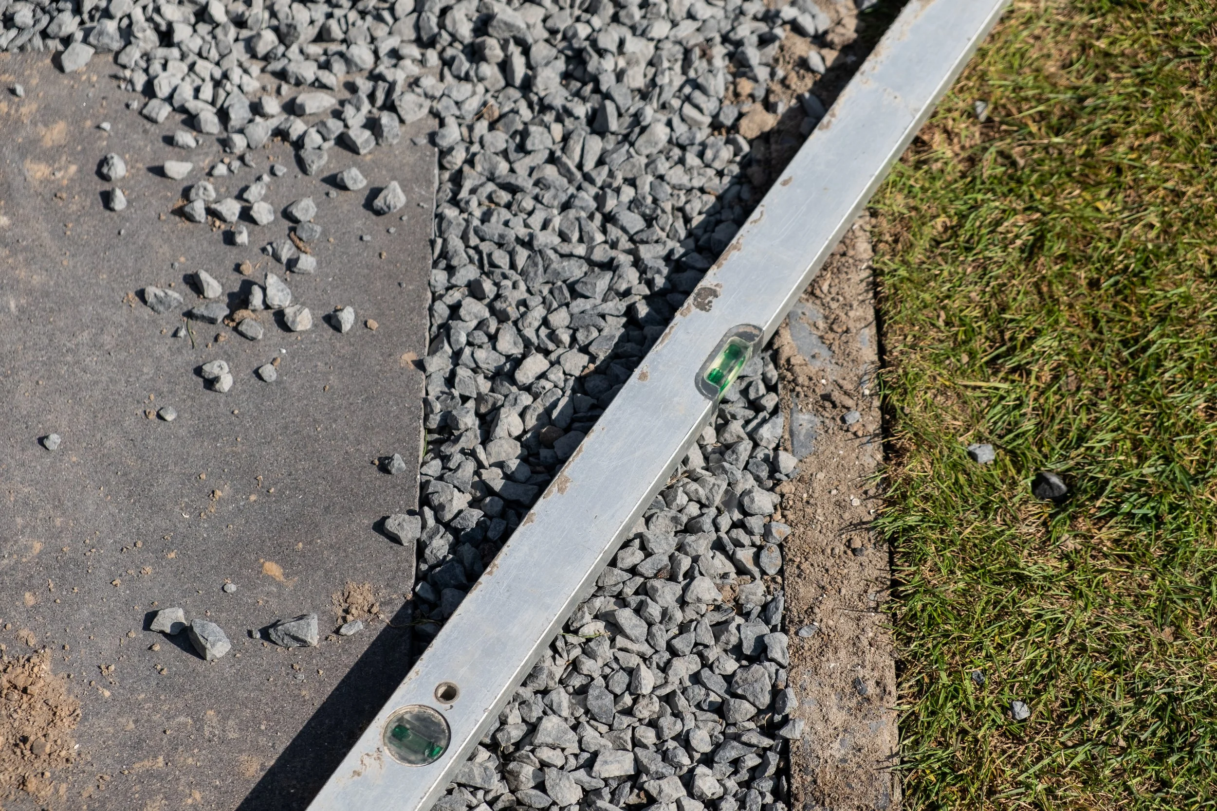 Close-up of a level tool placed along the edge of a newly laid concrete pavement and gravel border, with grass on the right side.