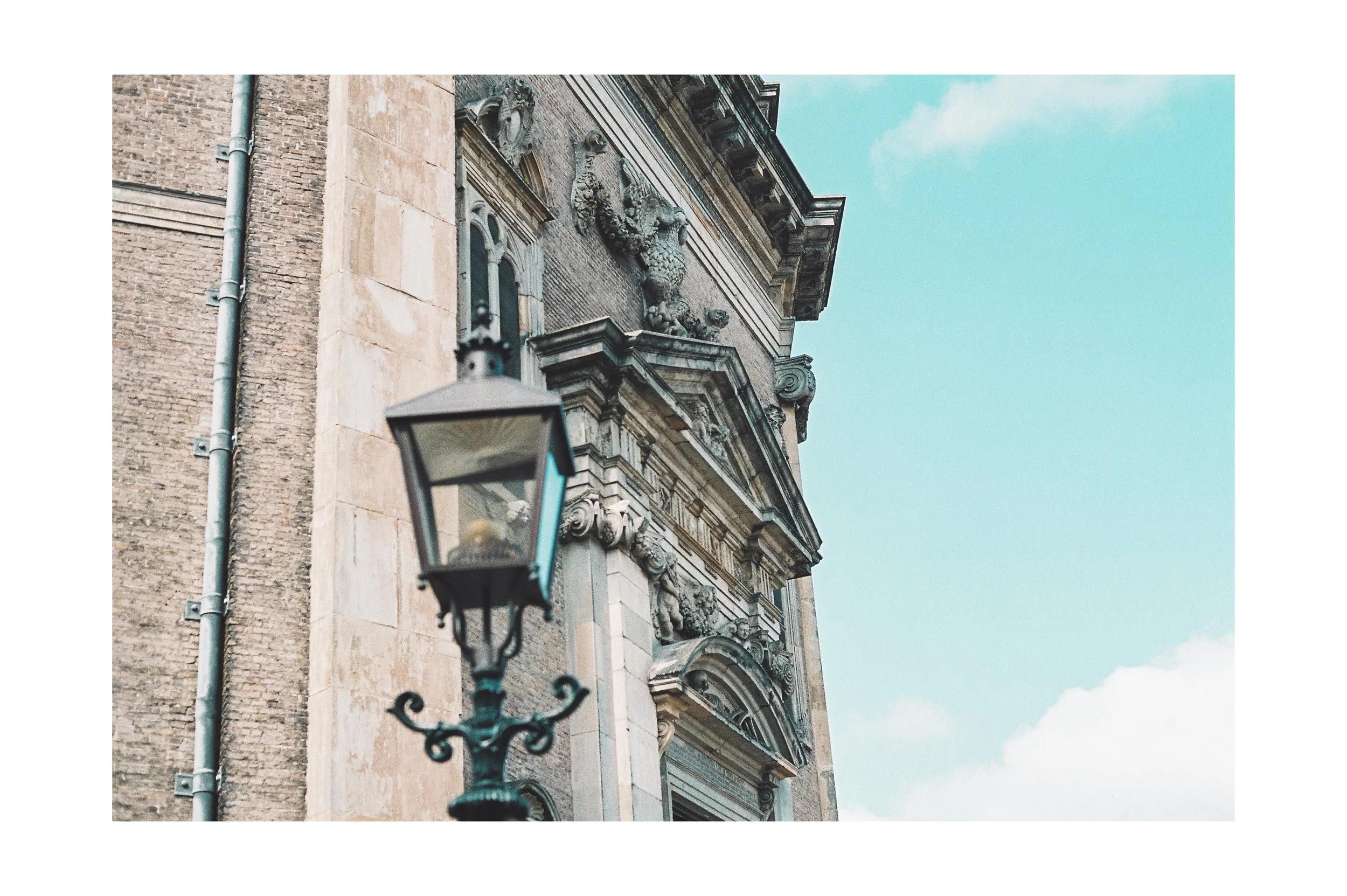Close-up of an ornate building facade with classical architectural details and sculptures, with an antique street lamp in the foreground against a blue sky.