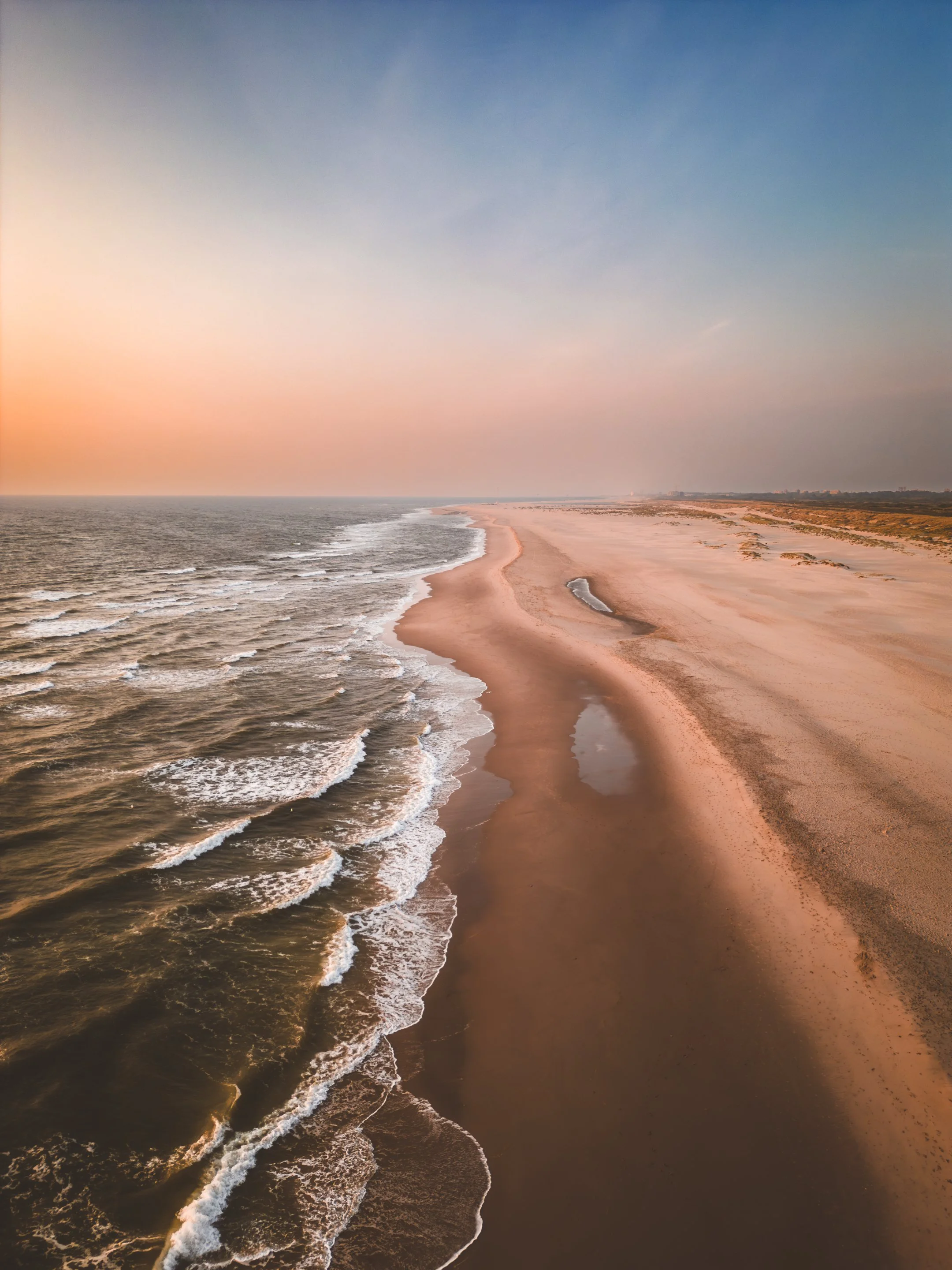 A wide view of a sandy beach with ocean waves on the left and a clear sky with soft pink and blue hues in the background.