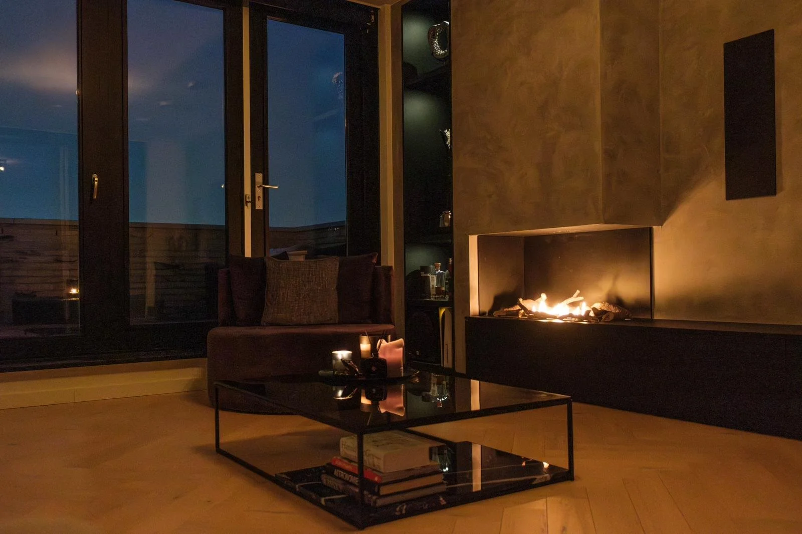 Cozy living room interior with a lit fireplace, a dark-colored sofa with pillows, a modern glass coffee table with books and candles, and large windows showing a nighttime view outside.