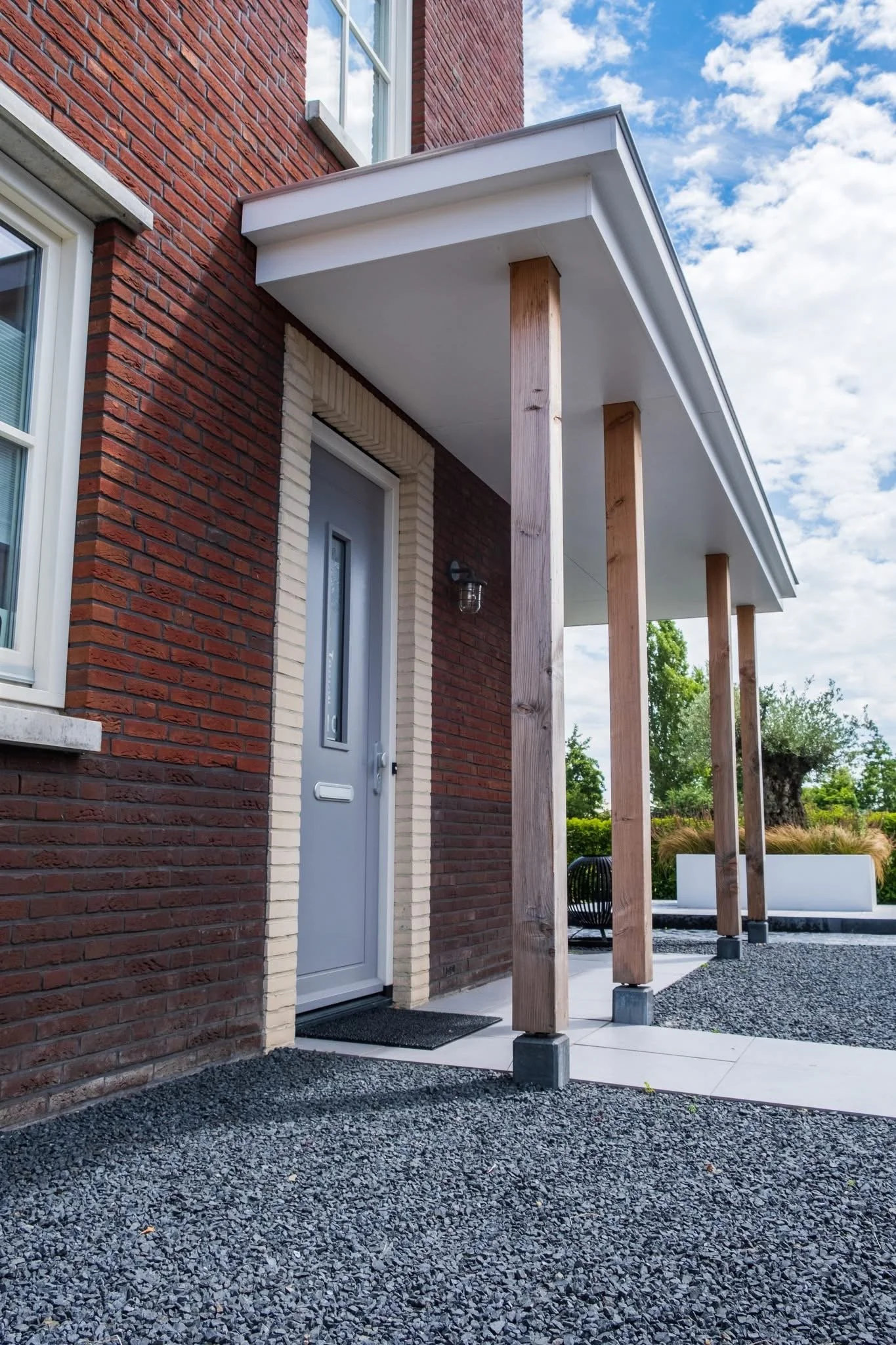 Exterior view of a modern house entrance with a door, brick walls, concrete and gravel ground, and wooden pillars supporting a white roof over the entrance.