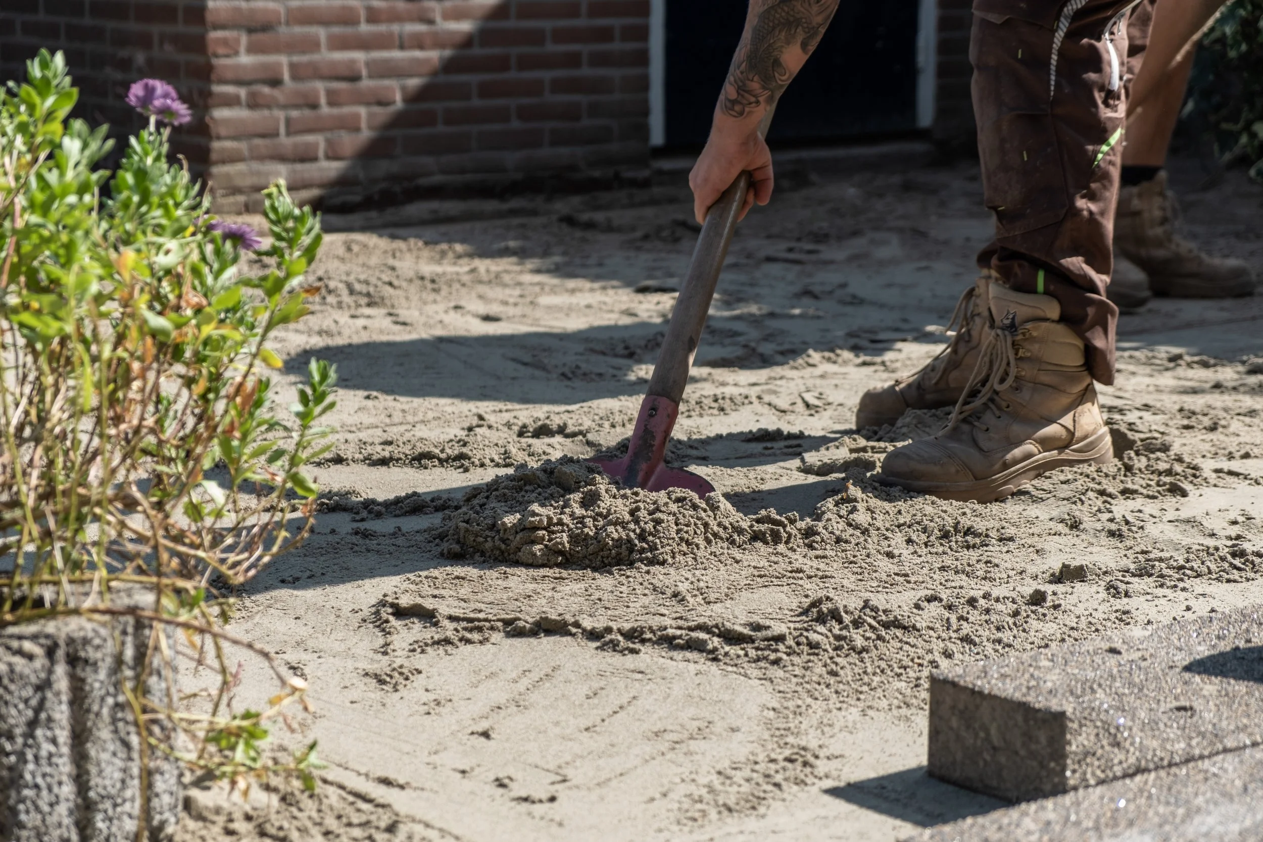 Person shoveling sand or dirt in a garden or construction area, wearing brown work boots and brown pants, with part of a brick wall and some plants visible in the background.