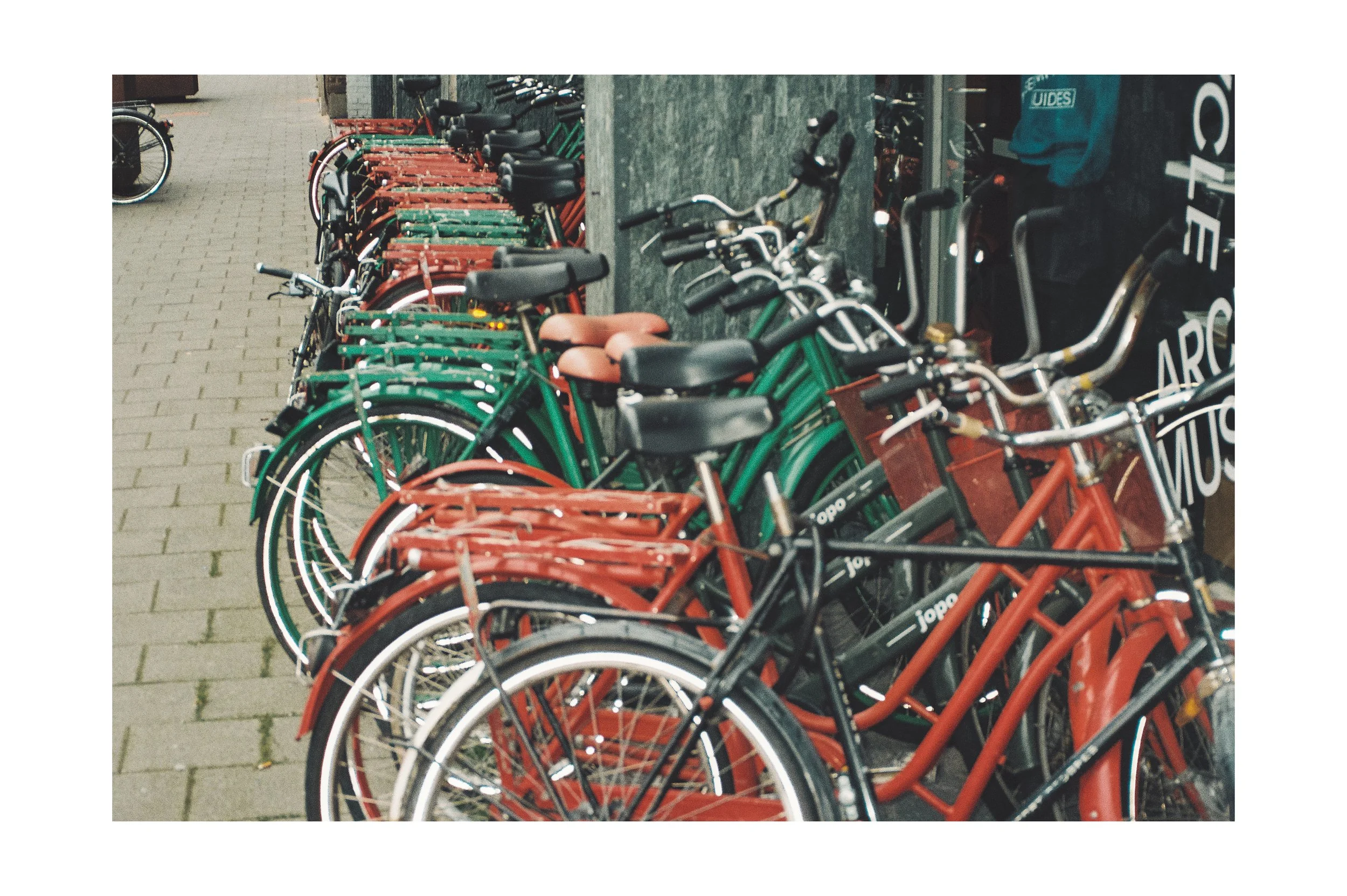 Row of green and red bicycles parked outside storefront, some with brown and black seats, sidewalk with brick pavers.
