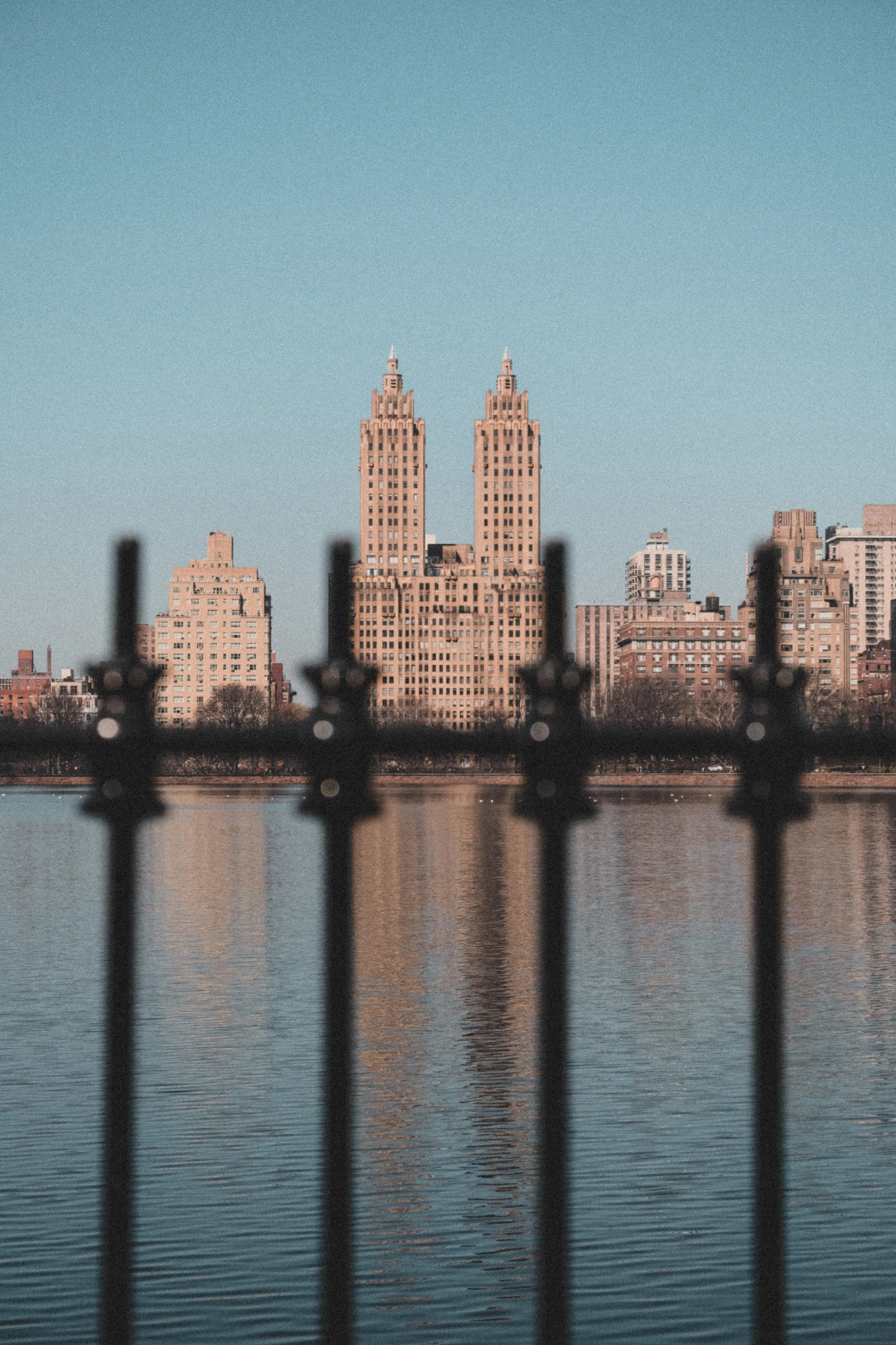 Two tall Art Deco style buildings viewed across a body of water, with a blurred black fence in the foreground and a clear blue sky above. NEW YORK CITY SKYLINE