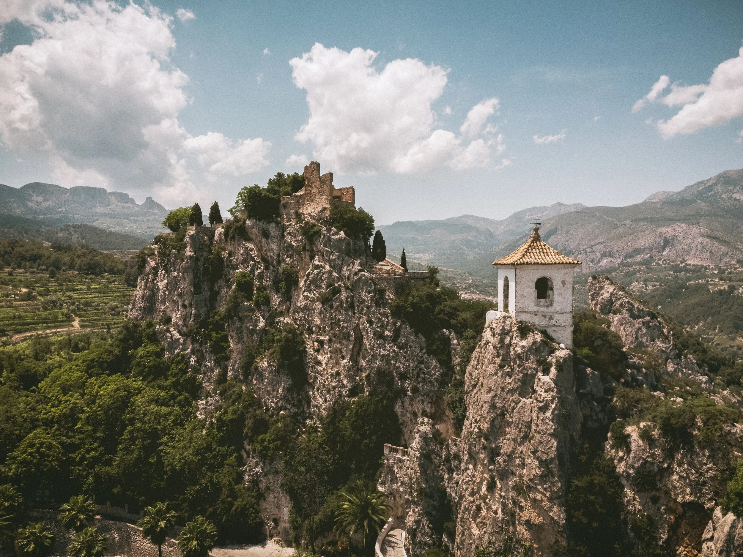 A castle ruins on a rocky hill with a white chapel on a rock formation in the foreground, set against a backdrop of mountains and a partly cloudy sky.