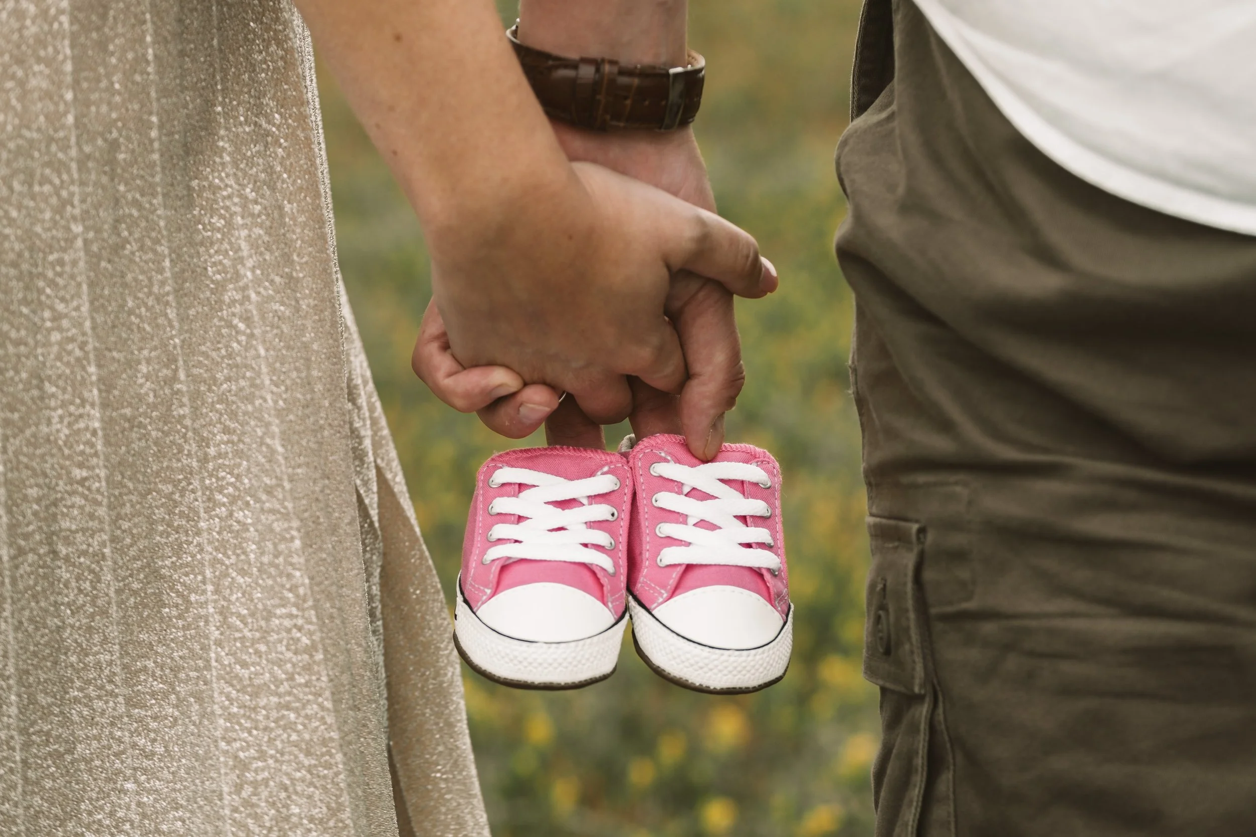 A couple holding tiny pink baby shoes between their hands, with a blurred outdoor background.