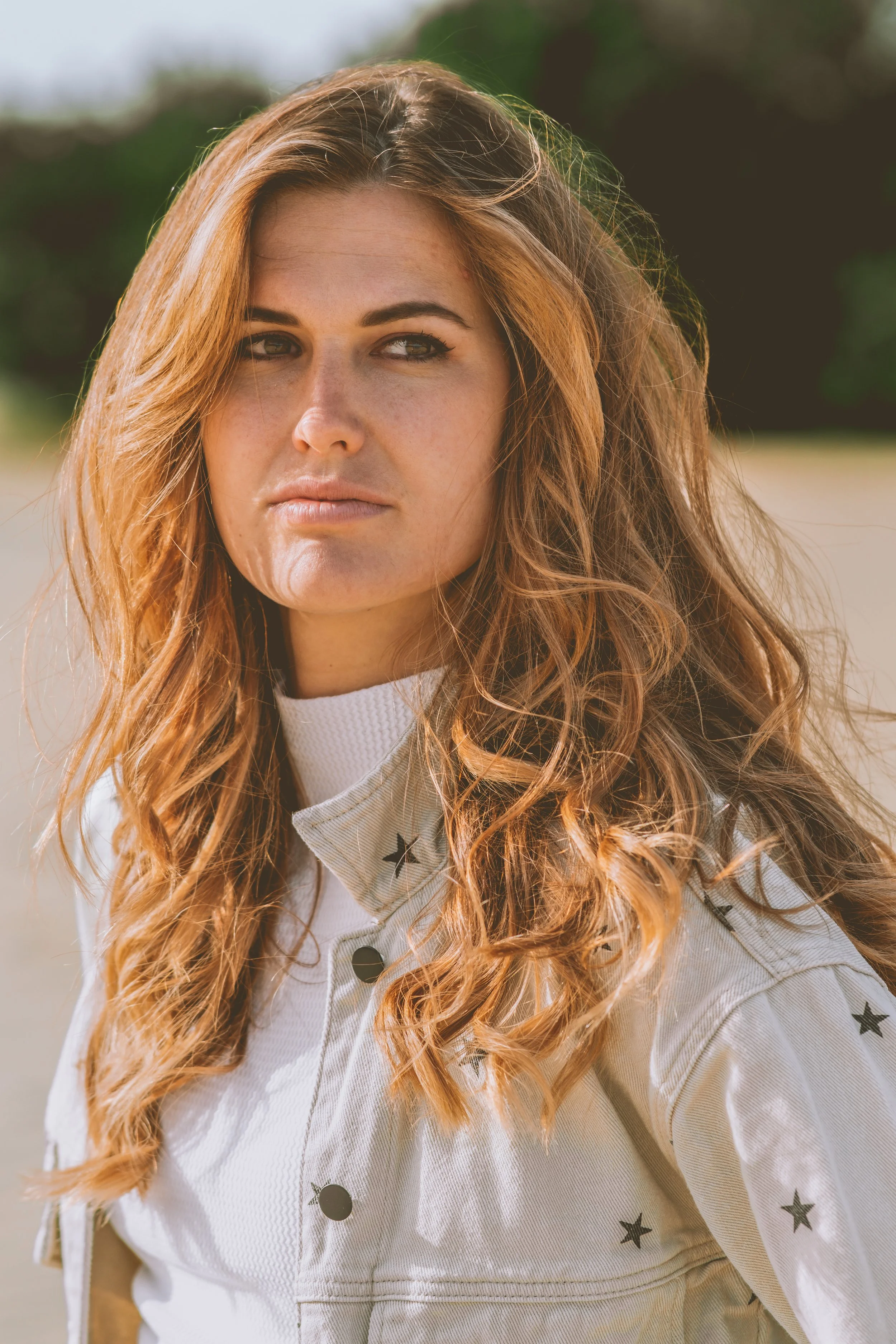 Close-up of a woman with long, wavy red hair outdoors, wearing a white jacket with black stars and black buttons.