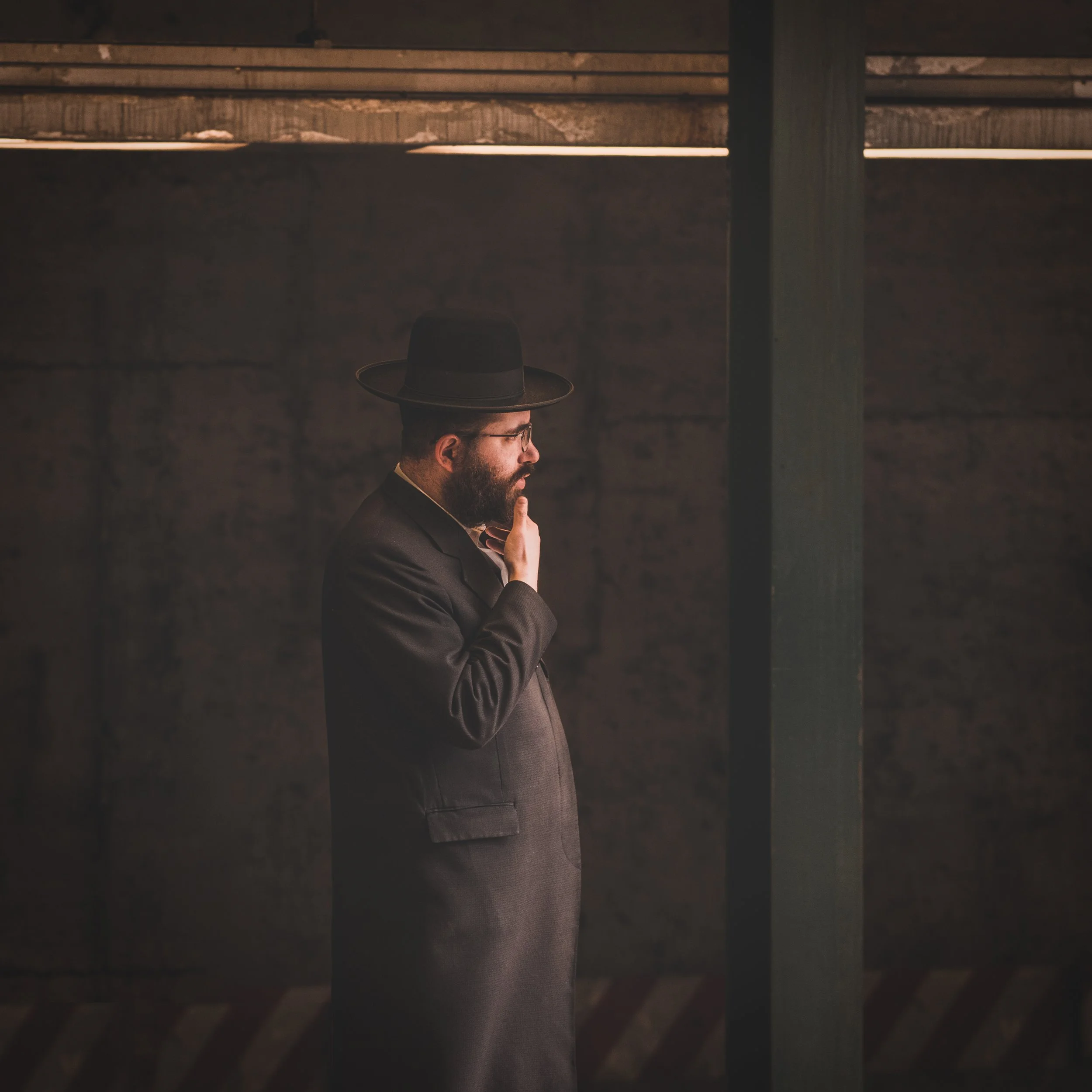 A man dressed in a black suit and hat, with glasses and a beard, standing sideways with his hand on his chin, in a dimly lit industrial setting.