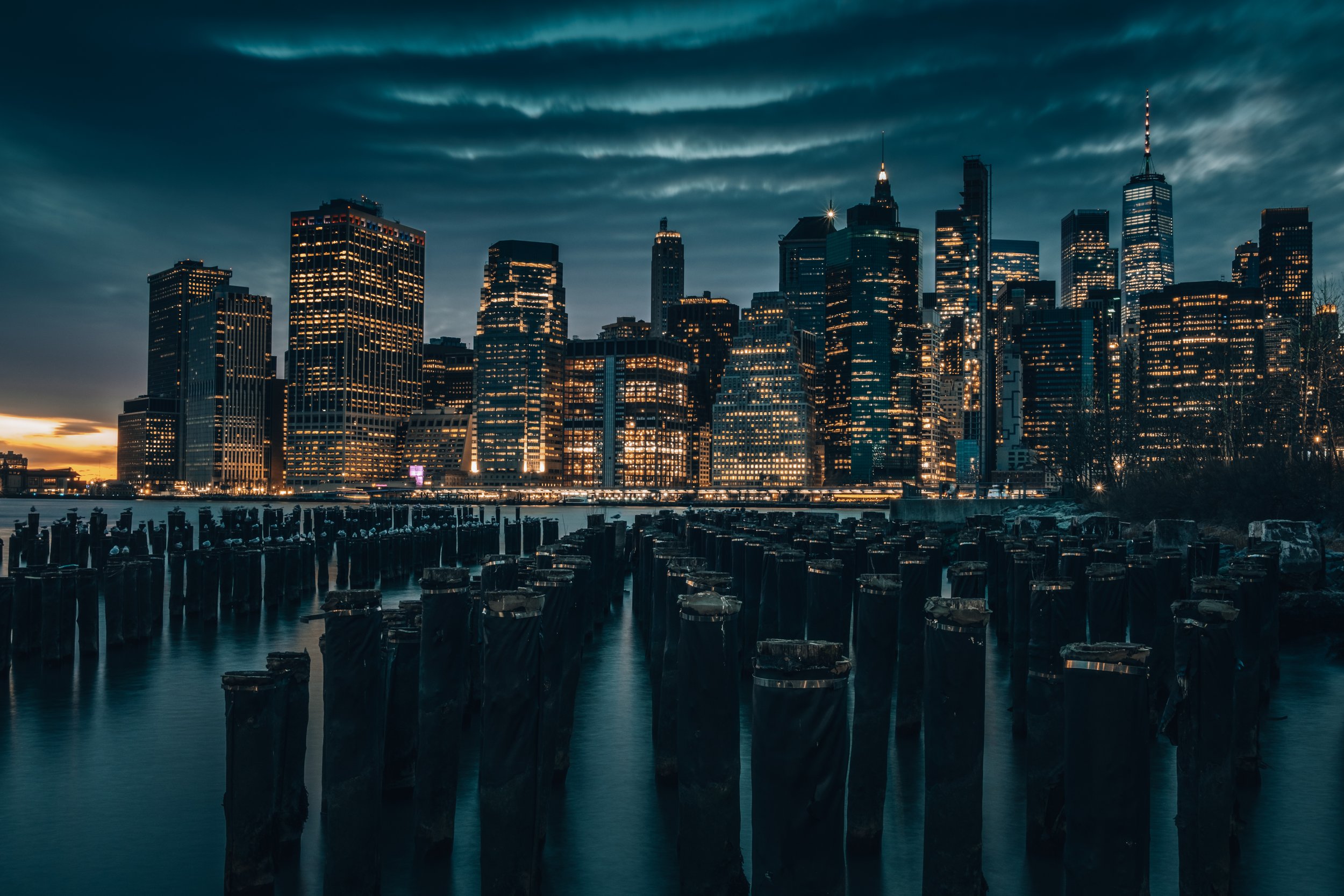 Nighttime view of a city skyline with tall illuminated buildings and a waterfront with old wooden pilings in the foreground.