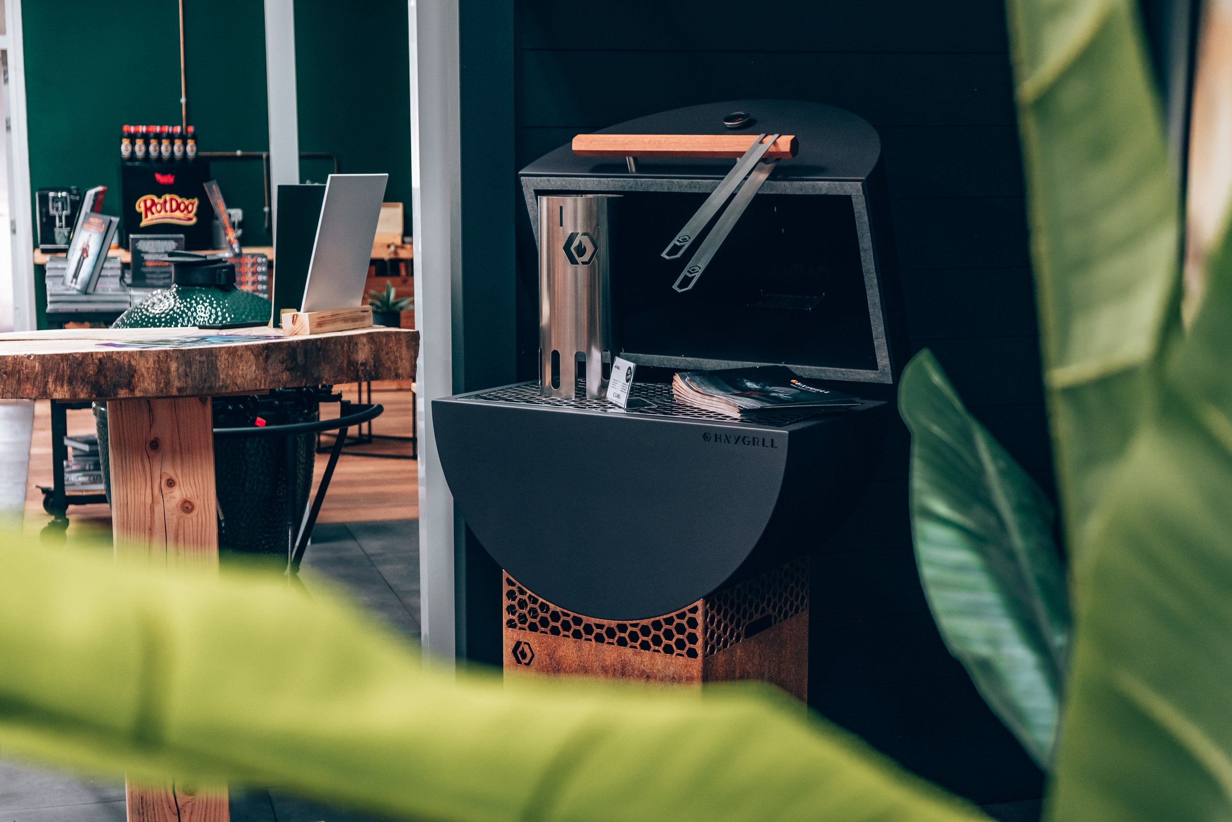 Indoor scene featuring a black and silver YGG Grill with wooden and metal components, surrounded by green leaves in the foreground.