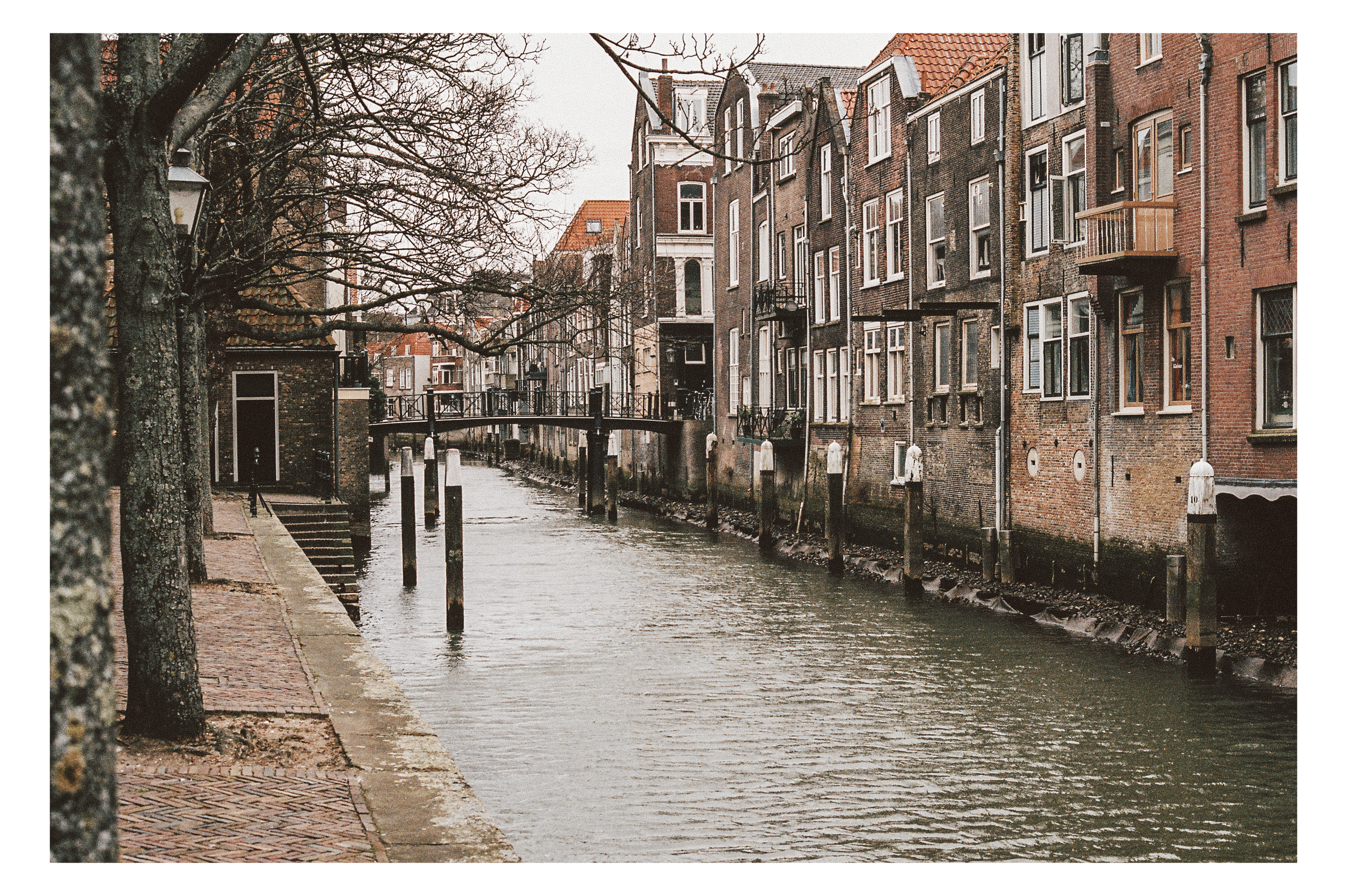 A canal with buildings and trees lining the sides, overcast sky, and calm water.