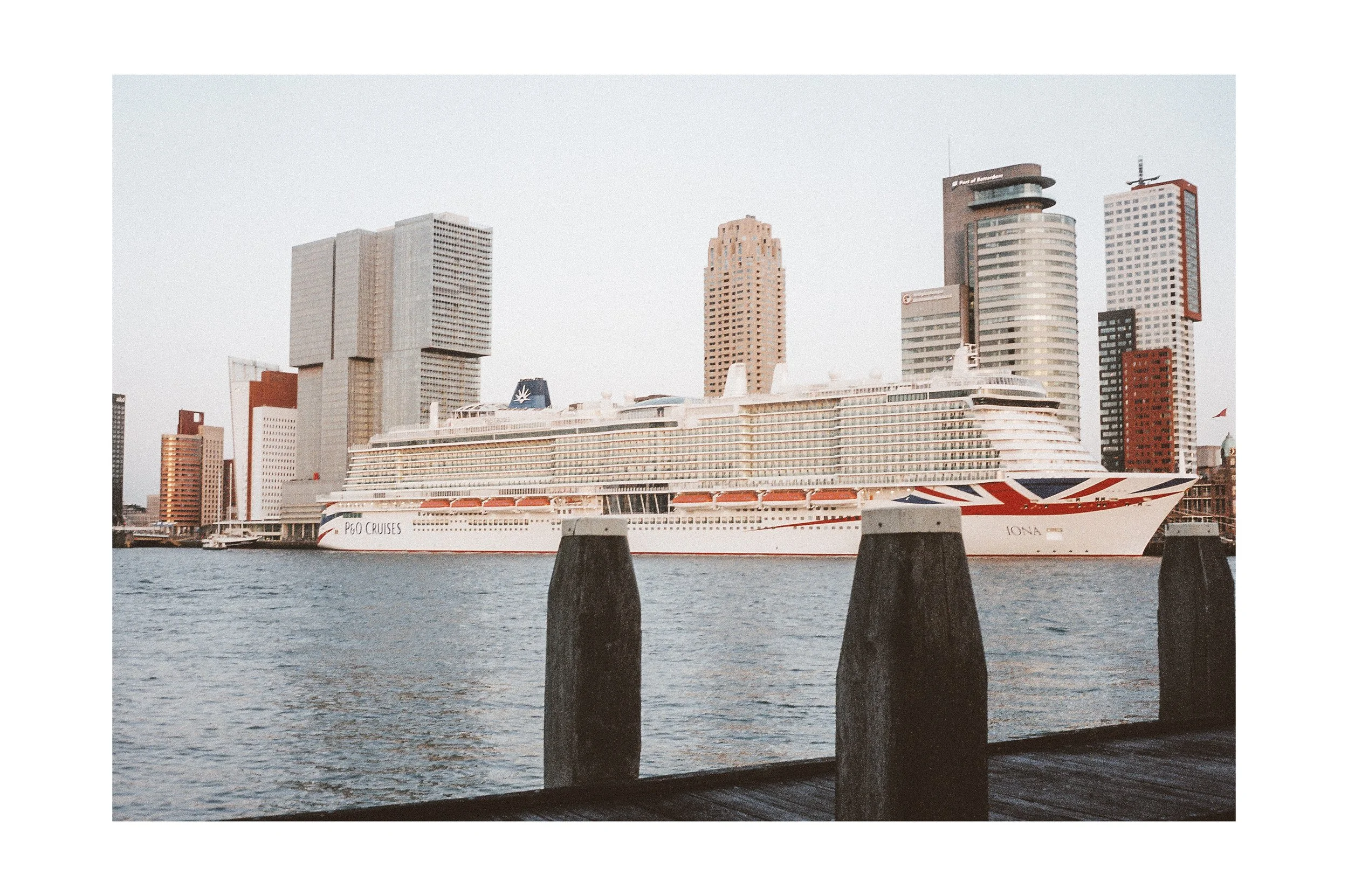 A large cruise ship docked in a city harbor with modern high-rise buildings in the background, near a wooden pier.