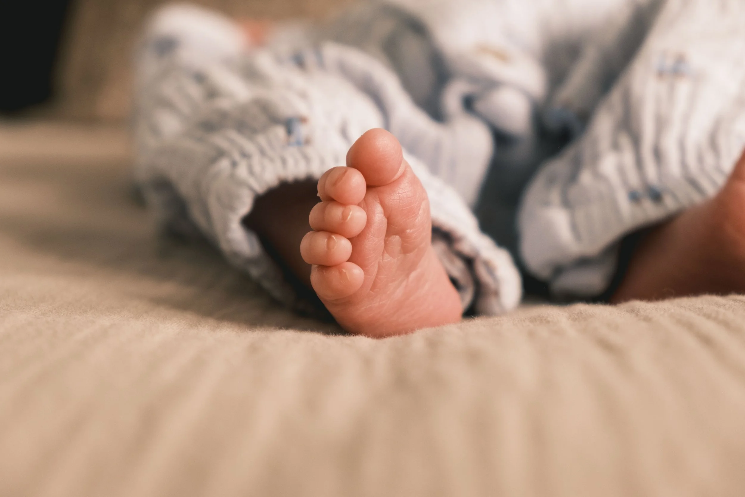 Close-up of a newborn baby's foot resting on a soft, beige surface. The baby is wearing light-colored, patterned pants.