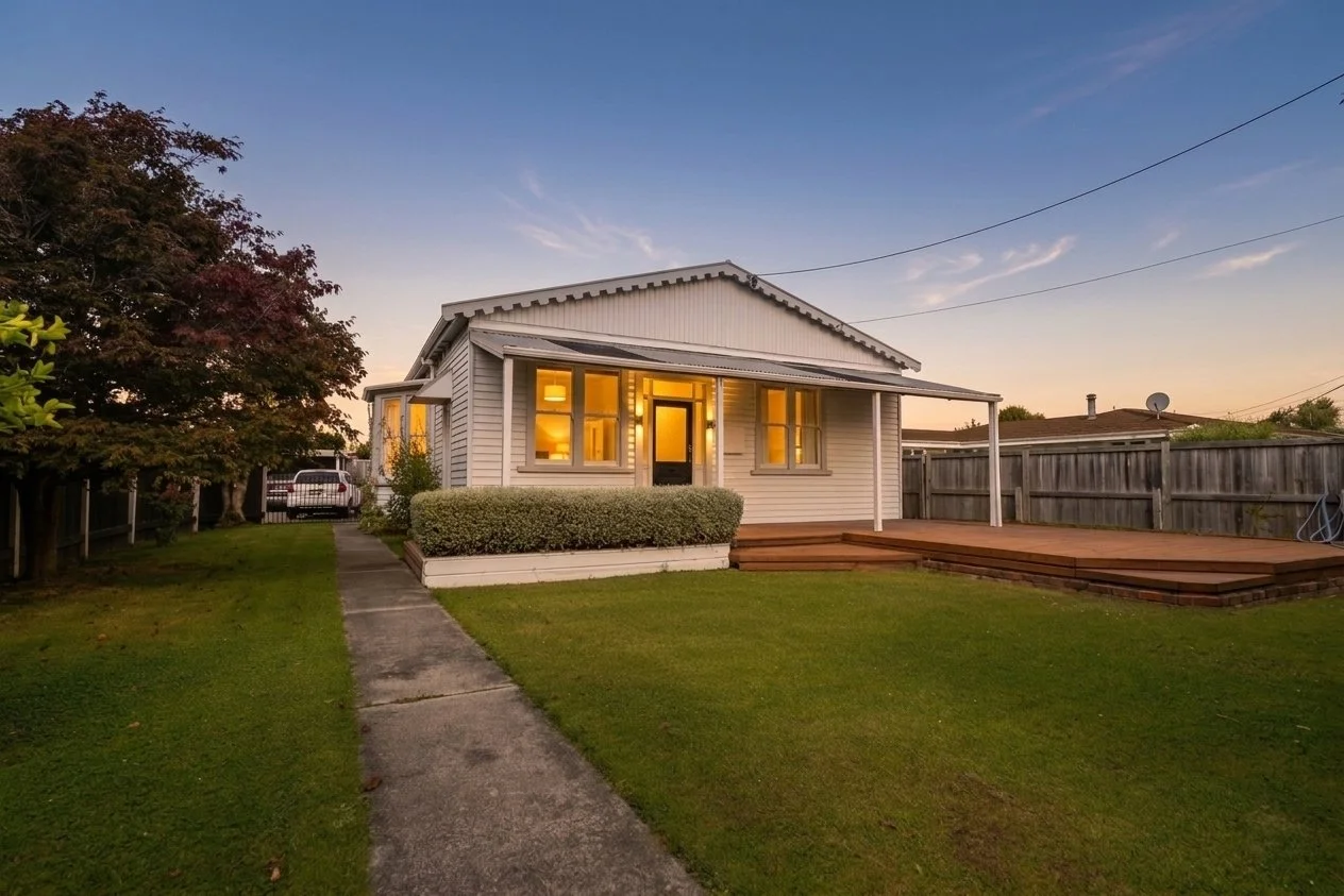 A cozy house with a white exterior, front porch, and wooden deck at sunset. The front yard has a concrete pathway, well-maintained lawn, and bushes. A tree is on the left, and a neighboring house and fence are visible in the background.