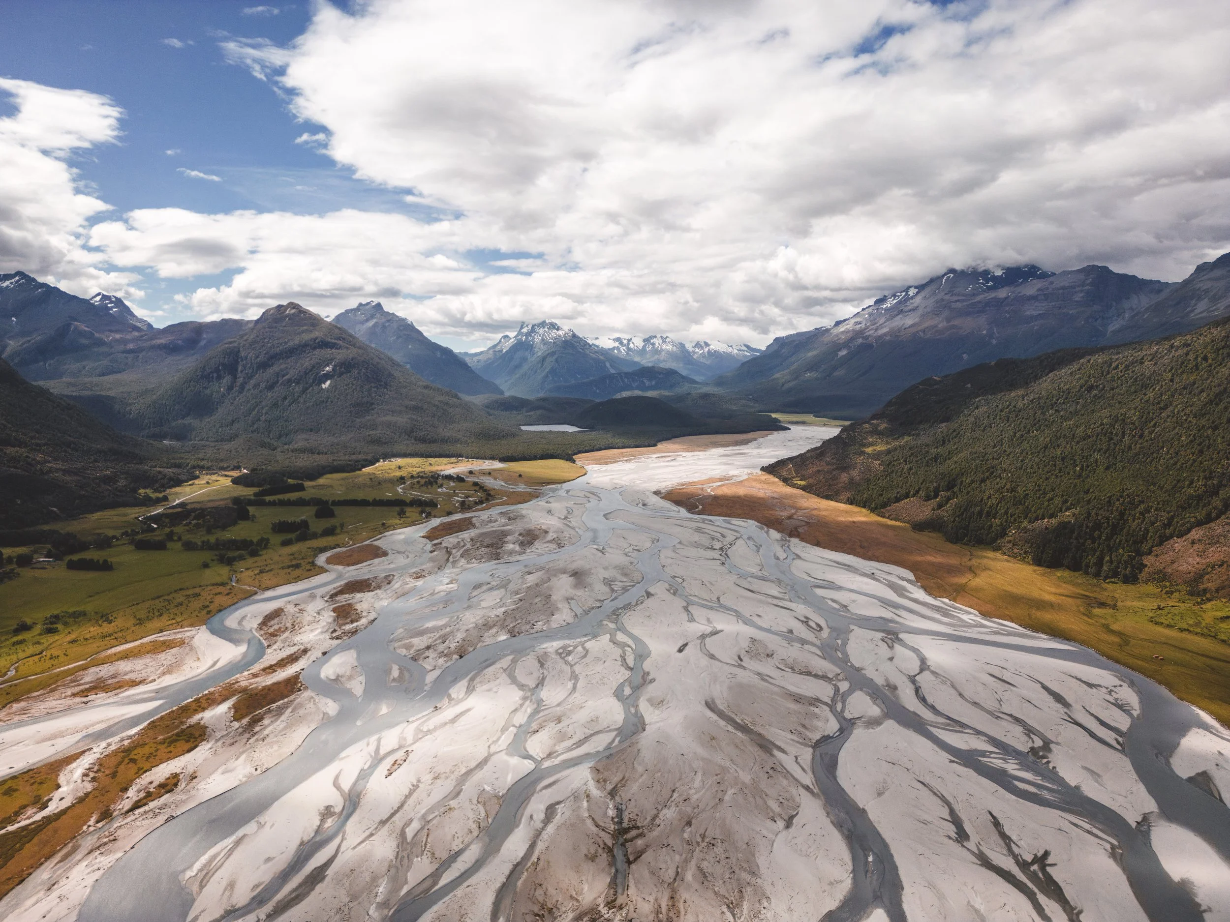 Aerial view of a river delta with multiple winding channels, surrounded by green fields, forested hills, and snow-capped mountains in the background under a cloudy sky.