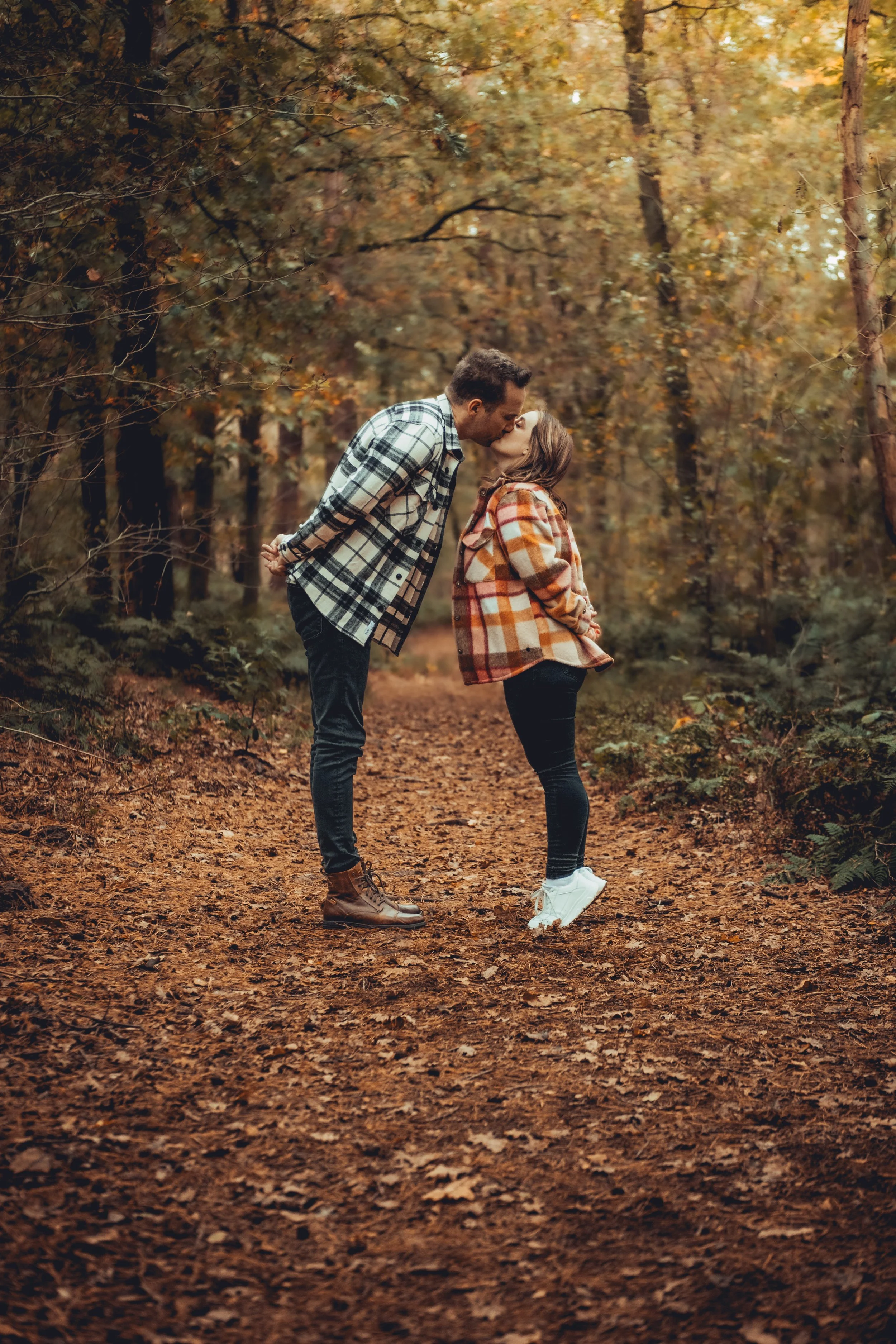 A couple sharing a kiss in a forest during autumn, surrounded by trees with fall foliage and fallen leaves on the ground.