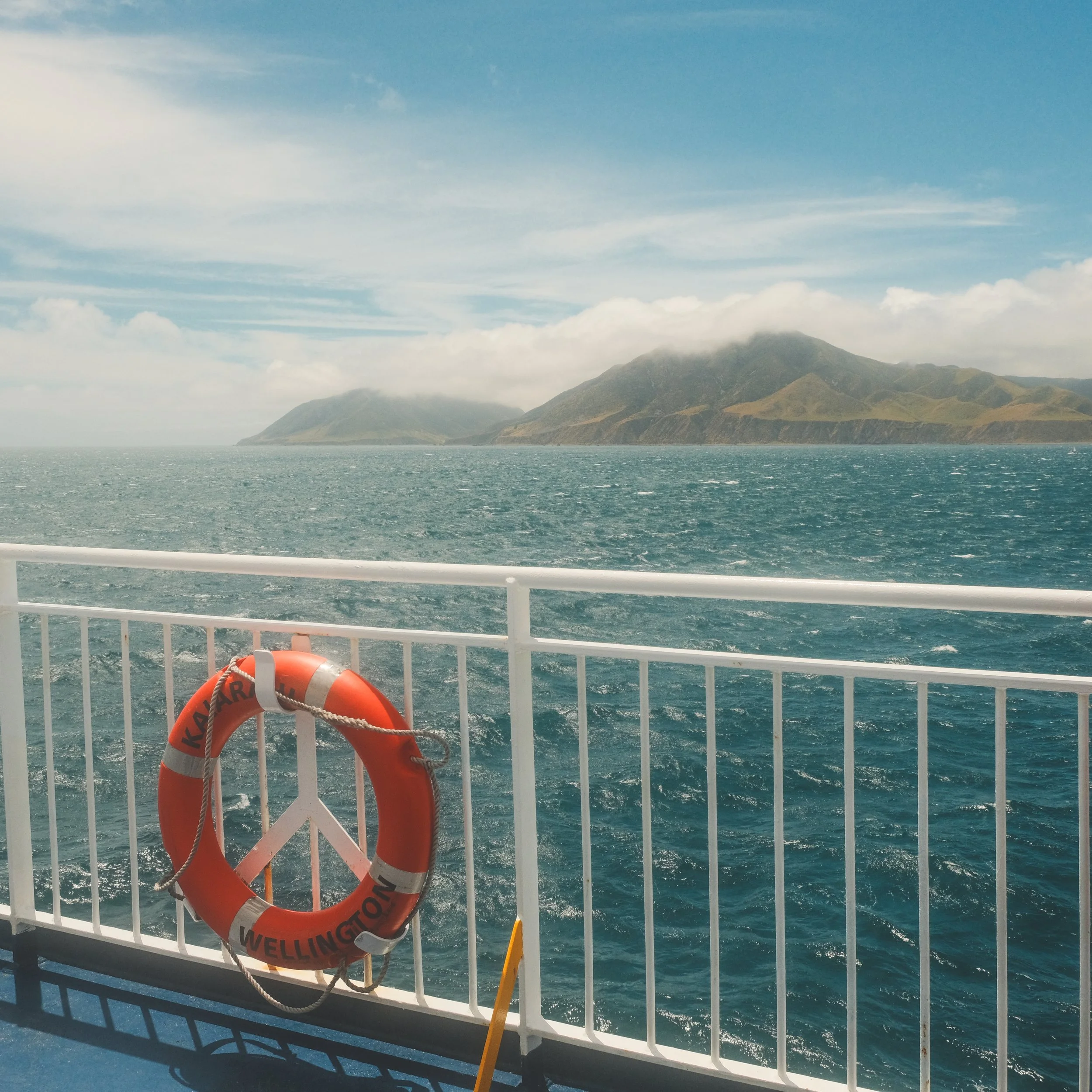 View from a boat showing the ocean, distant islands with mountains, a white safety railing, and an orange life preserver marked 'KARAKA WELLINGTON'.