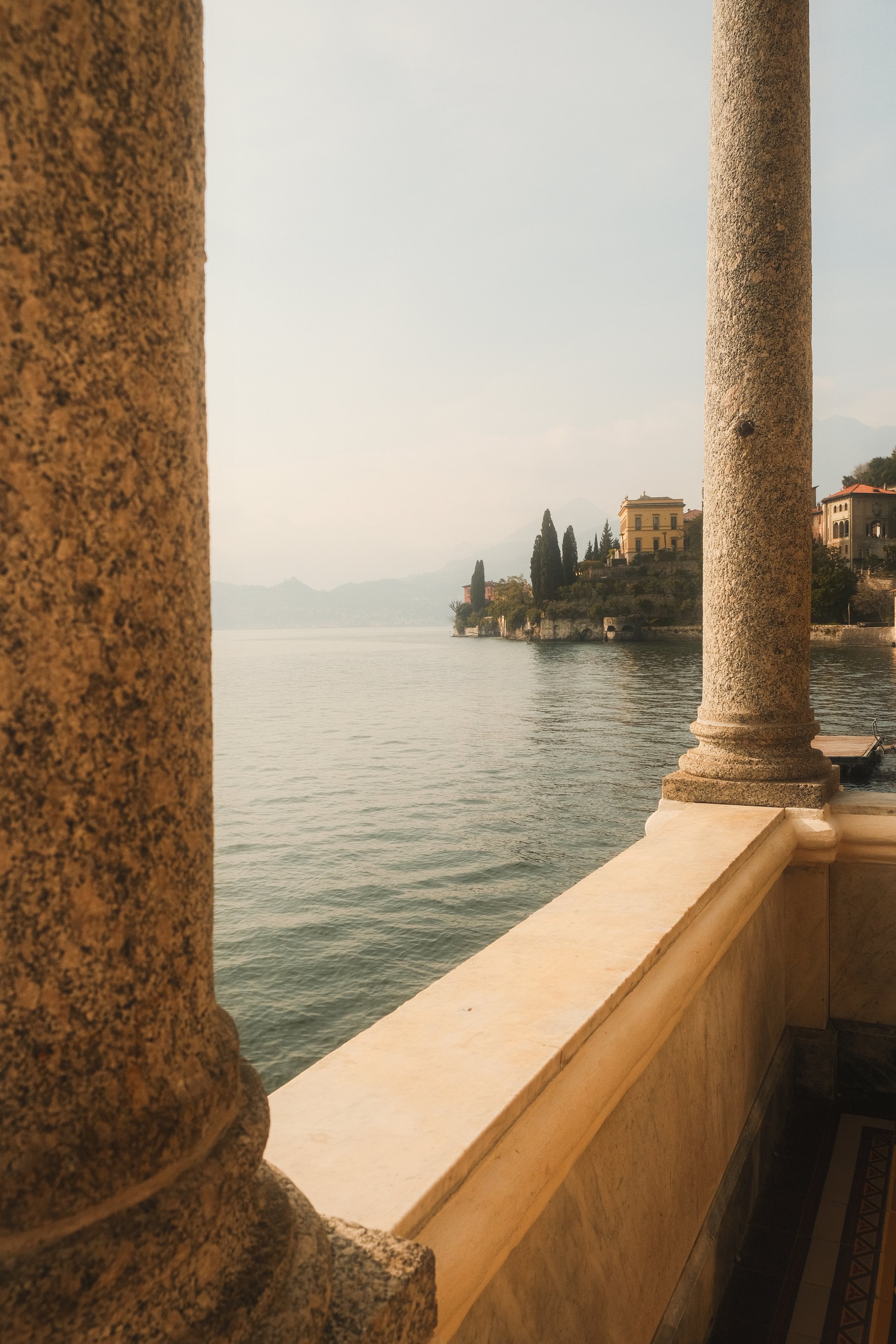 View of a lake seen from a balcony with stone columns, with houses and trees on a hill across the water, and misty mountains in the background.