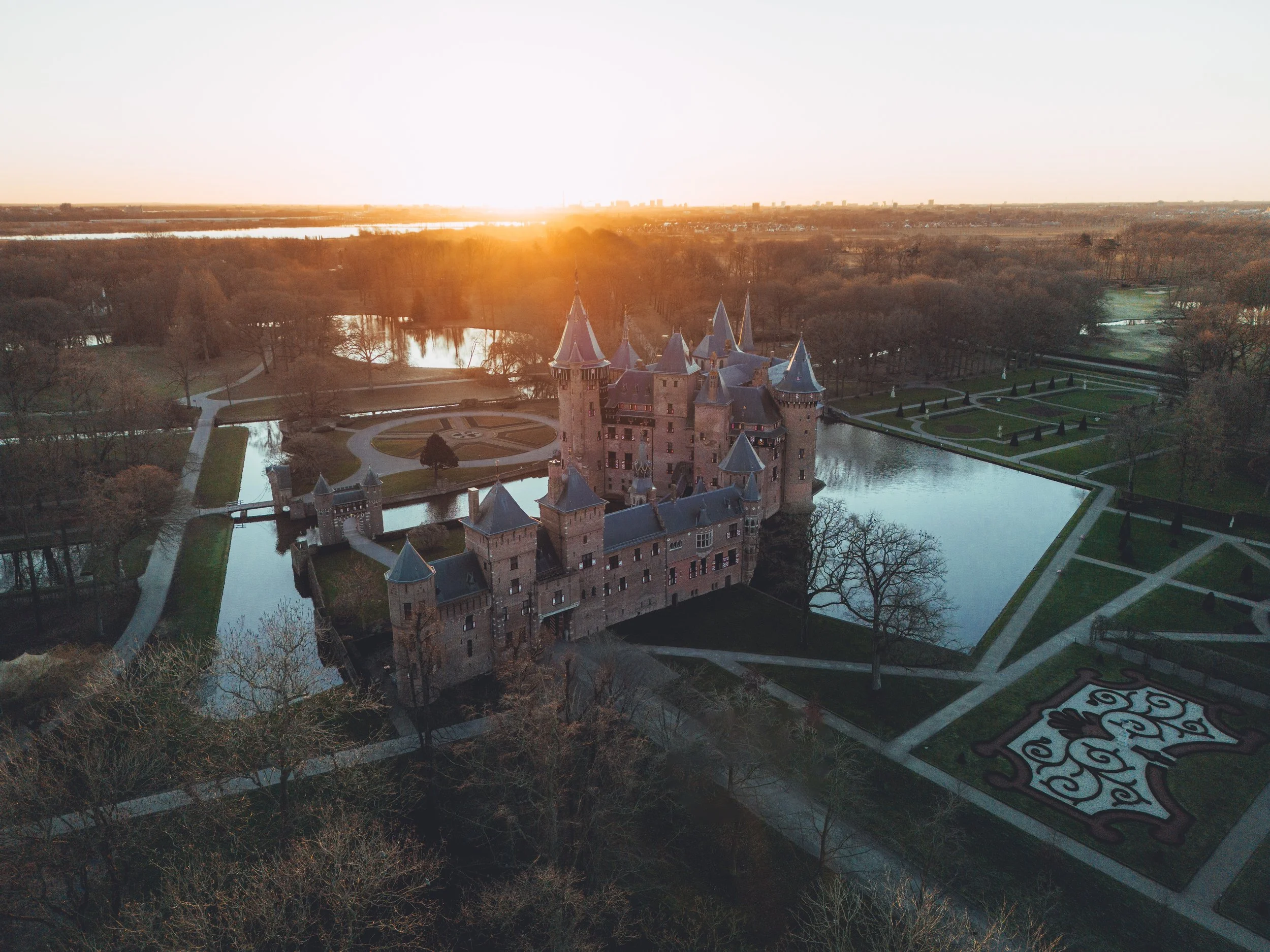 Aerial view of a castle surrounded by water with a park and walking paths at sunset.