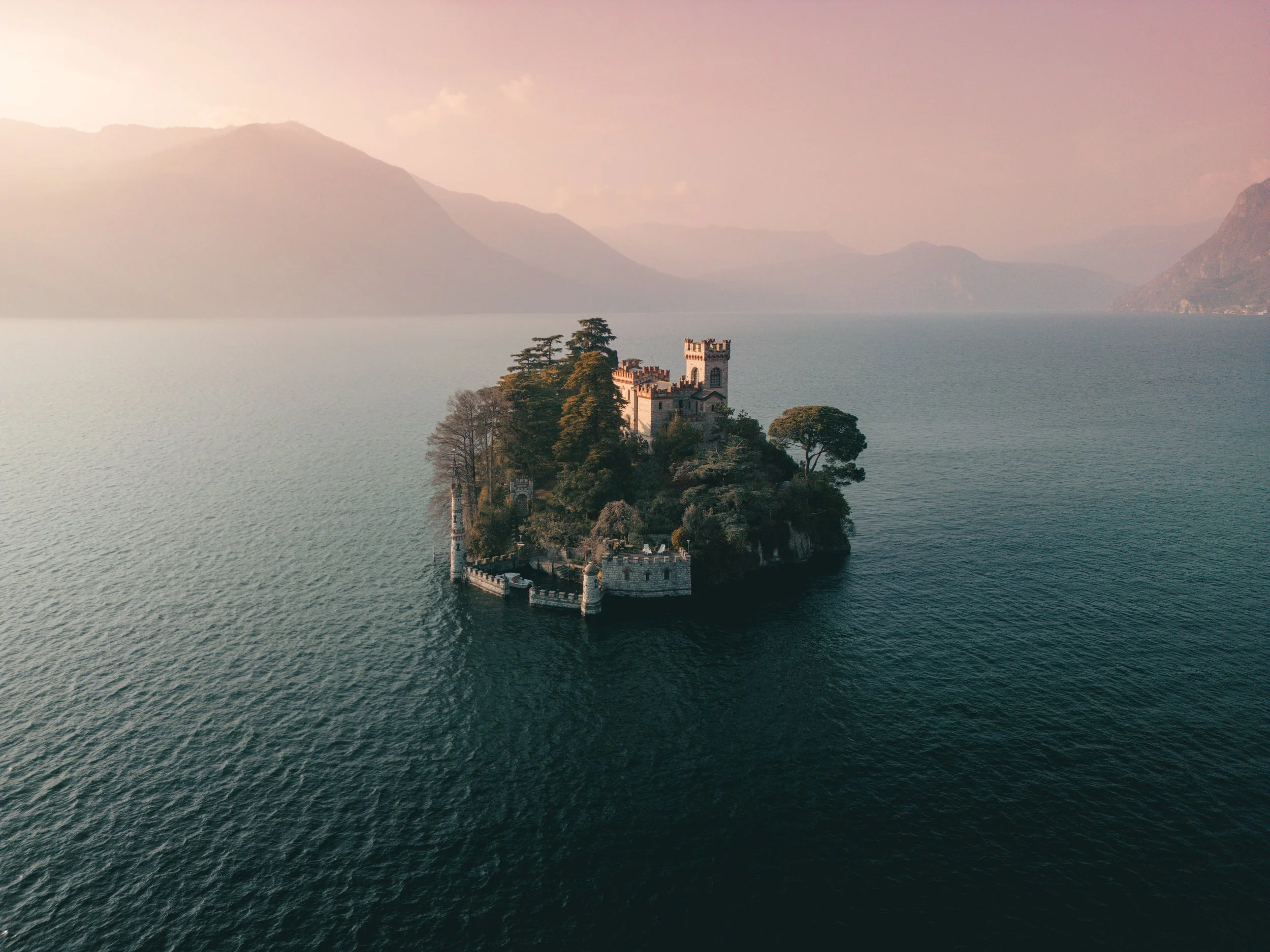 A small island with a castle and trees surrounded by a large body of water, with mountains in the background at sunset.
