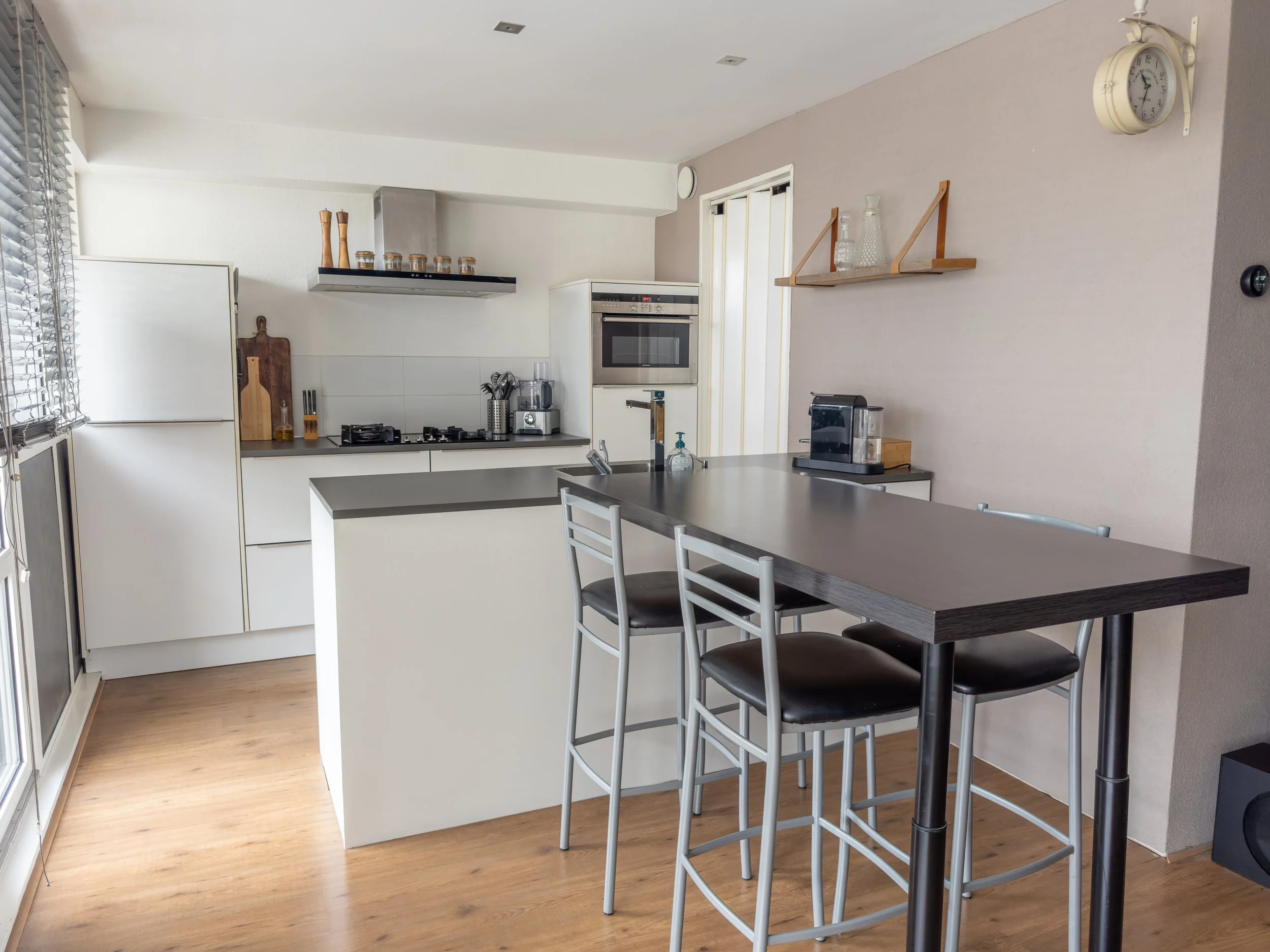Modern kitchen with white cabinets, gray countertops, and a kitchen island with three black-and-metal bar stools, hardwood floors, and small appliances on the counters.