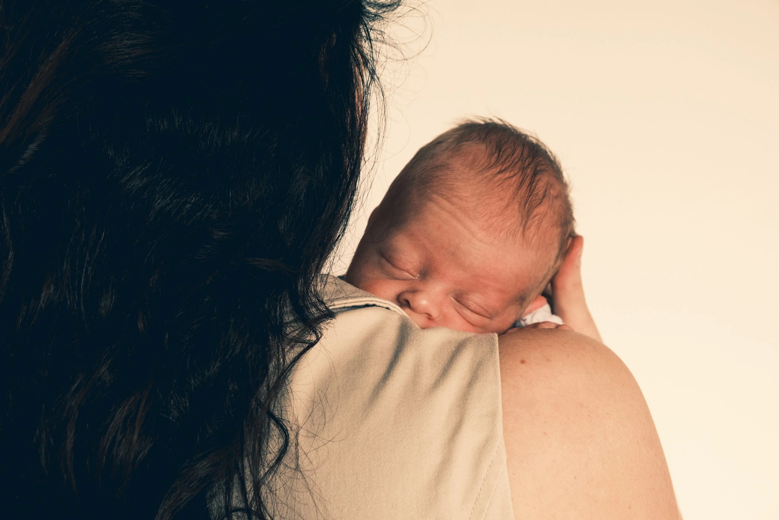 Close-up of a sleeping newborn baby resting on a woman's shoulder, with the woman's dark hair partially visible.