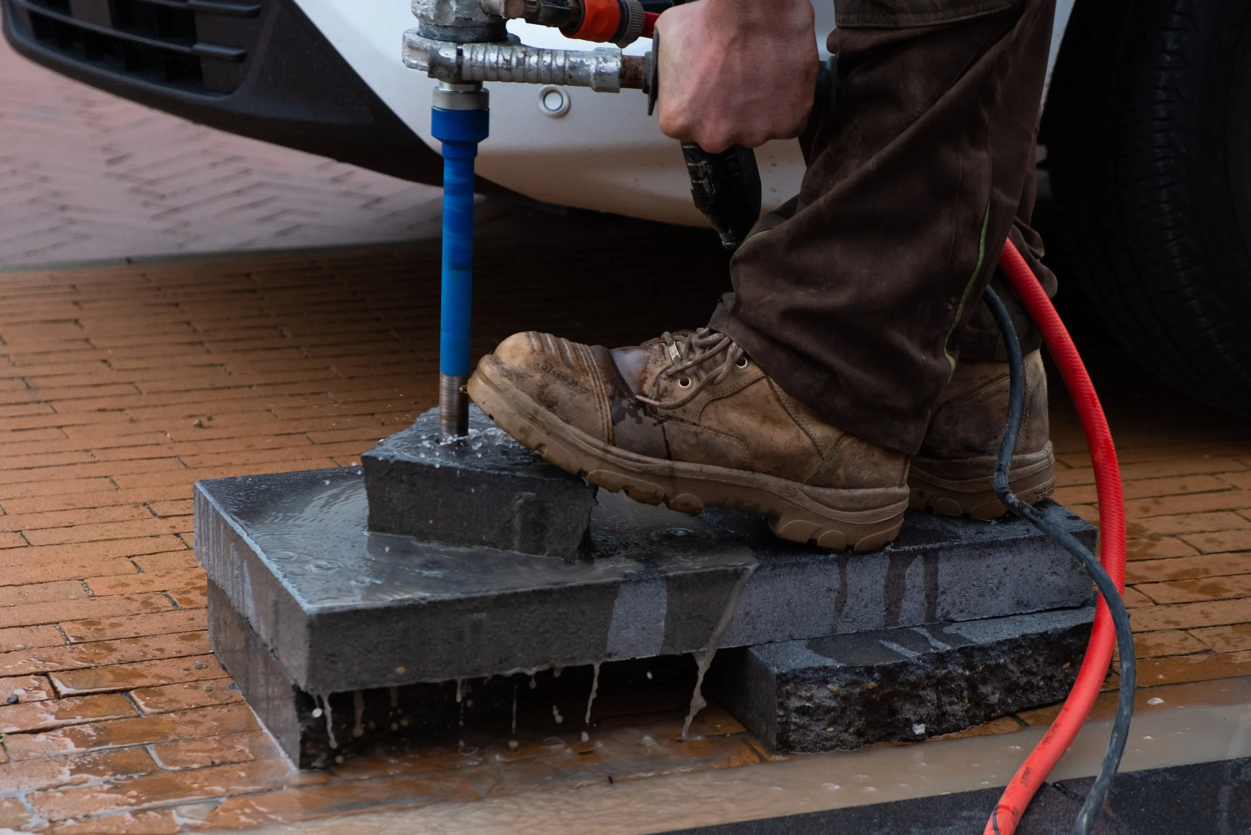 Person in brown work boots and brown pants using a jackhammer to break concrete on a brick surface.