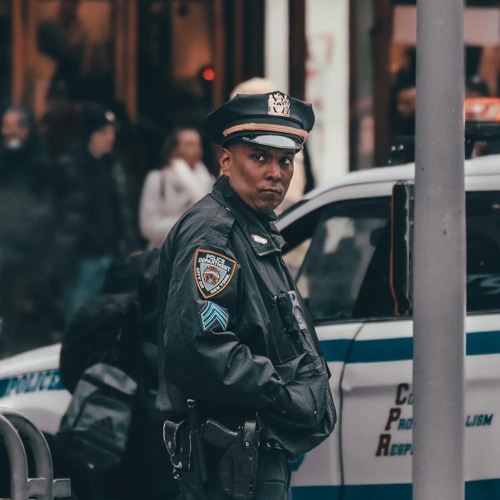 A police officer in uniform with a police badge and patches, standing on a city street near a police car.