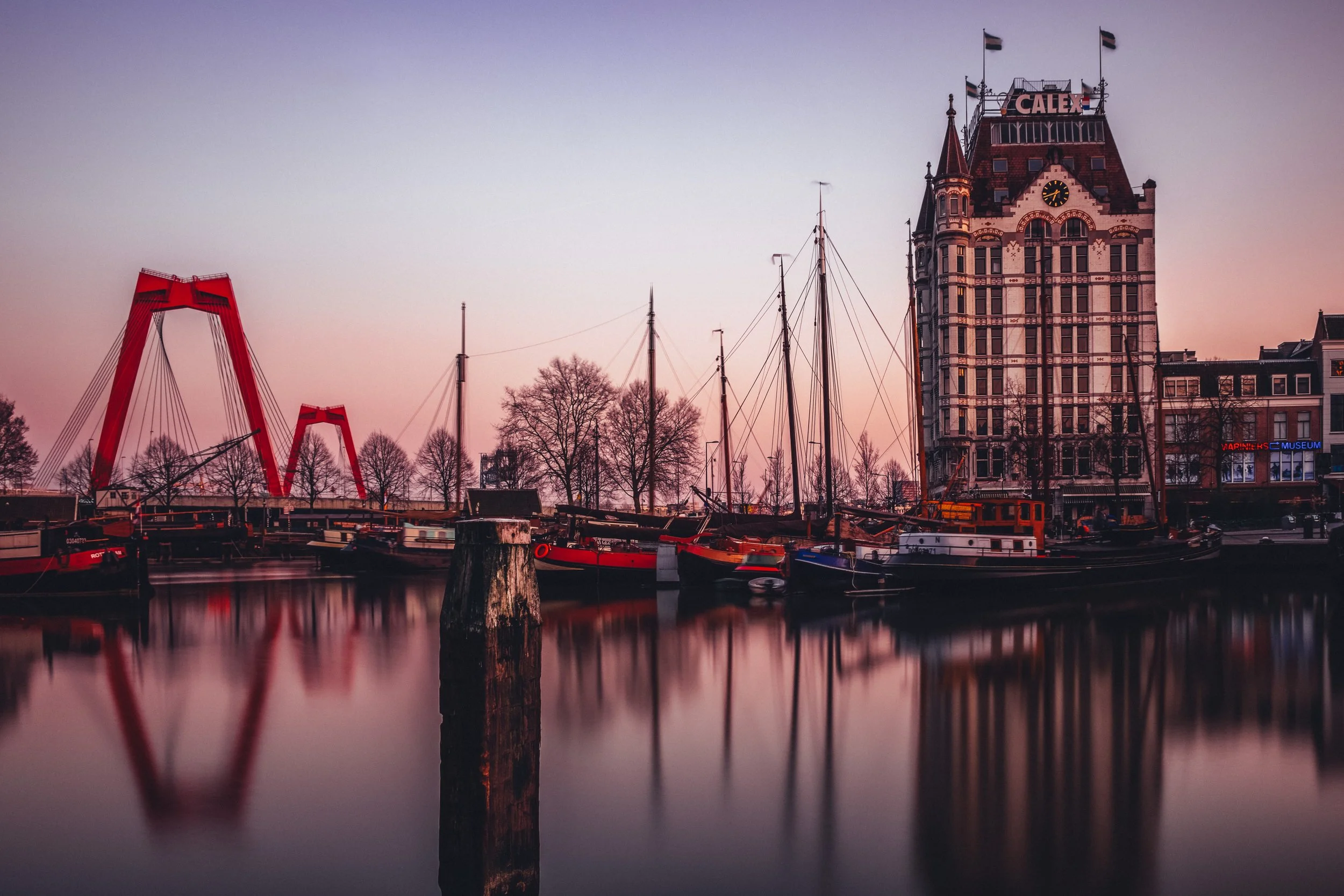 A waterfront scene at dusk featuring several boats docked along the pier, a tall historic building with a clock and the sign 'CALIX' on top, and a modern red bridge with twin towers in the background. The water reflects the boats and buildings.