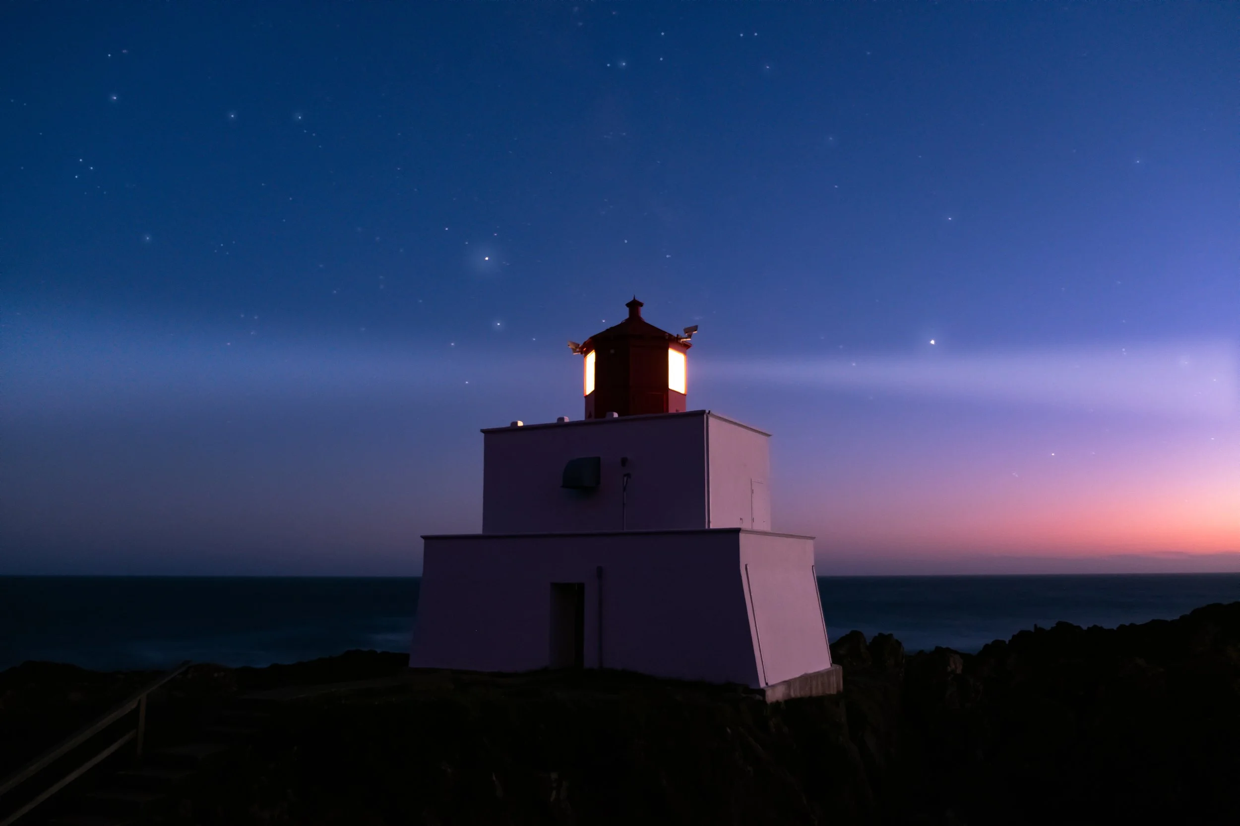 A lighthouse at dusk with a starry sky in the background and ocean waves below.