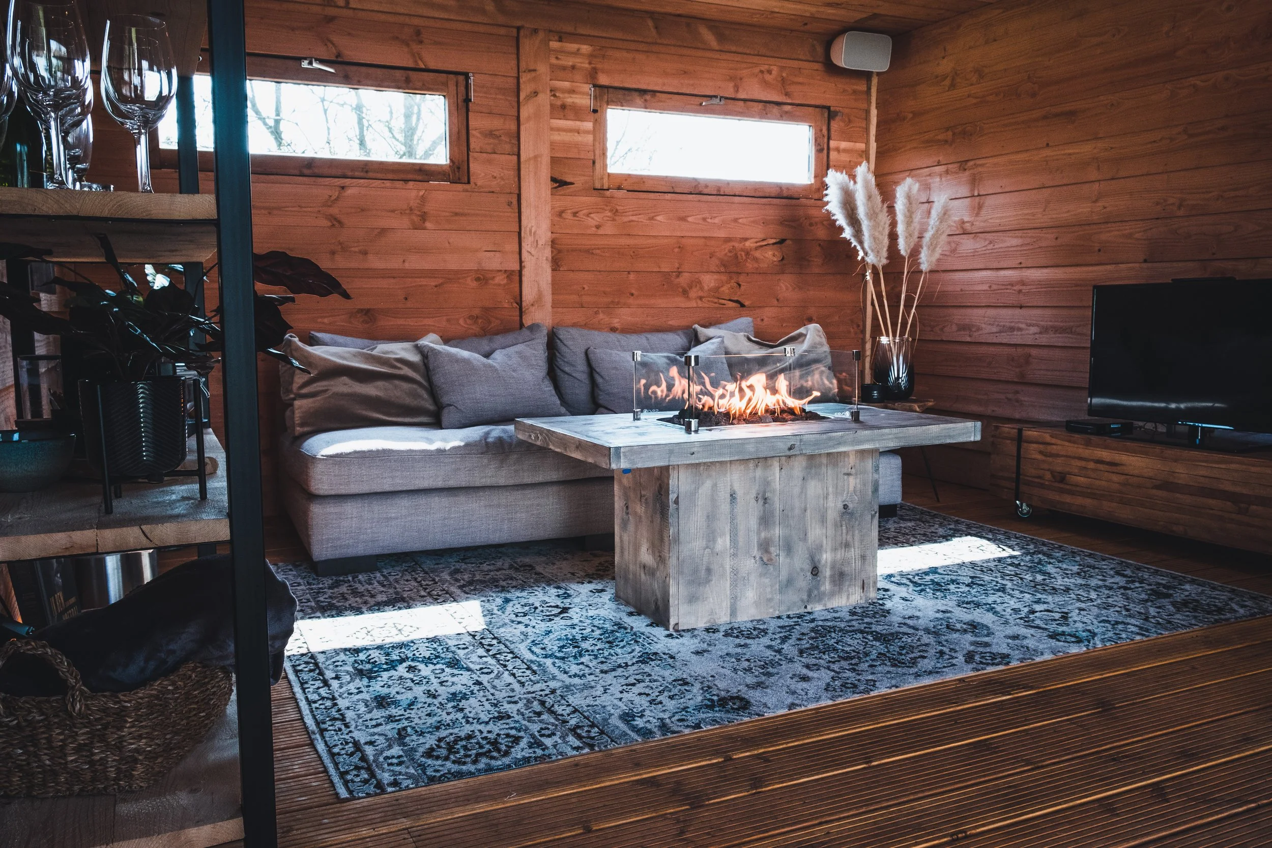 Living room with wooden walls, a gray sofa with beige and gray pillows, a modern fire table on a wooden stand, a patterned rug, a flat-screen TV, a wicker basket, and a black vase with pampas grass near a large TV.