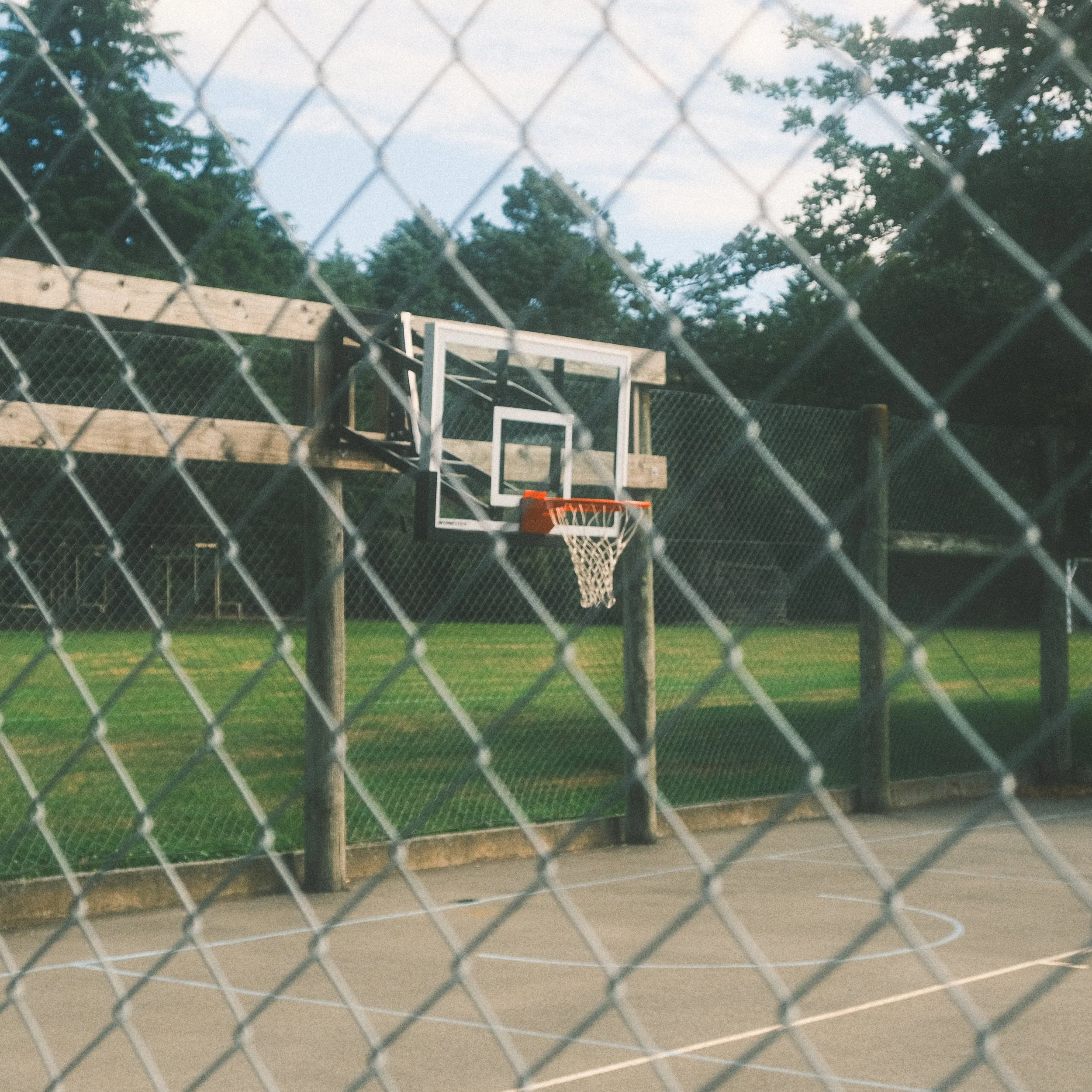 An outdoor basketball court with a hoop and backboard, surrounded by a chain-link fence, during evening light.