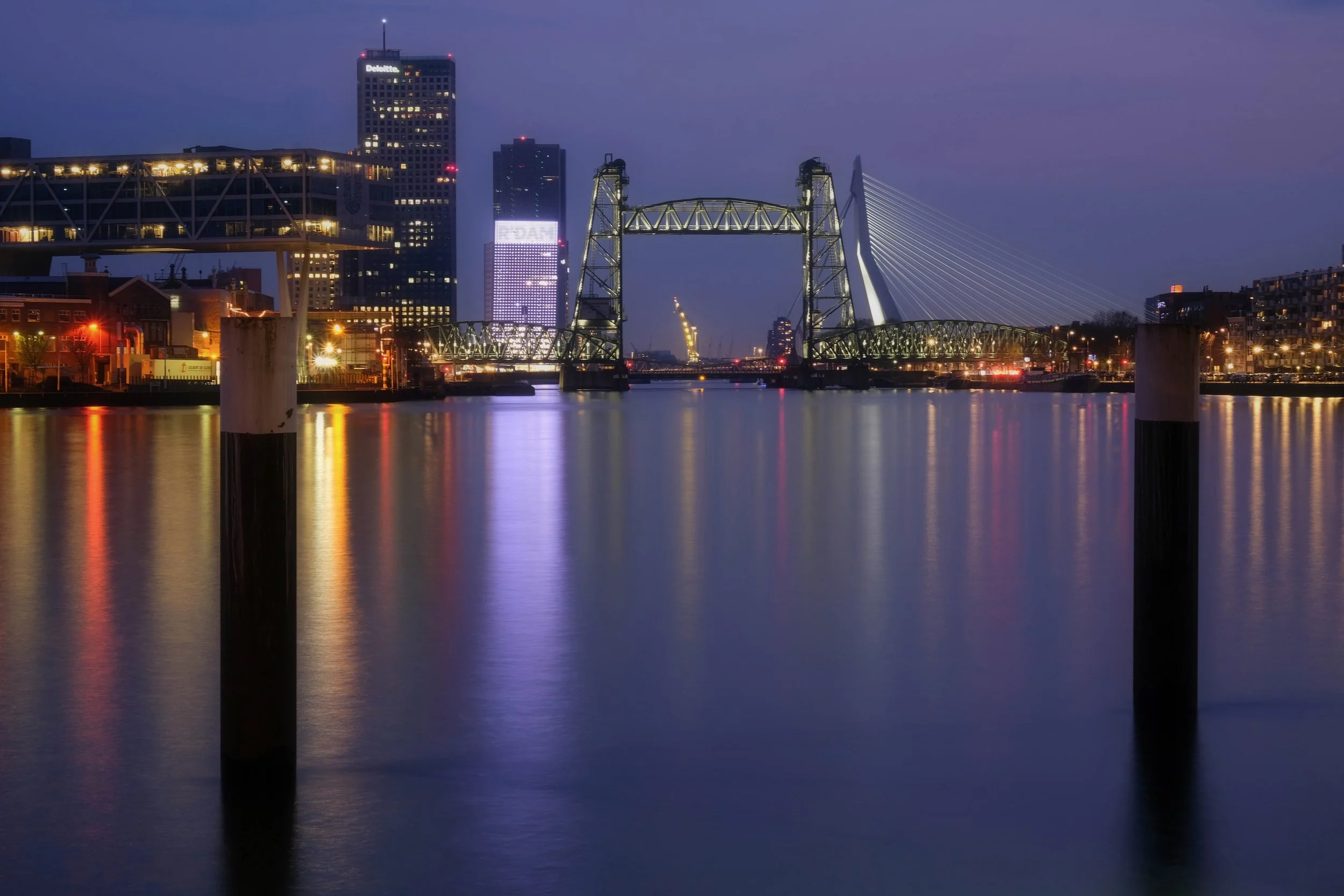 Nighttime cityscape of a river with illuminated buildings, a bridge, and a modern cable-stayed bridge in the background.