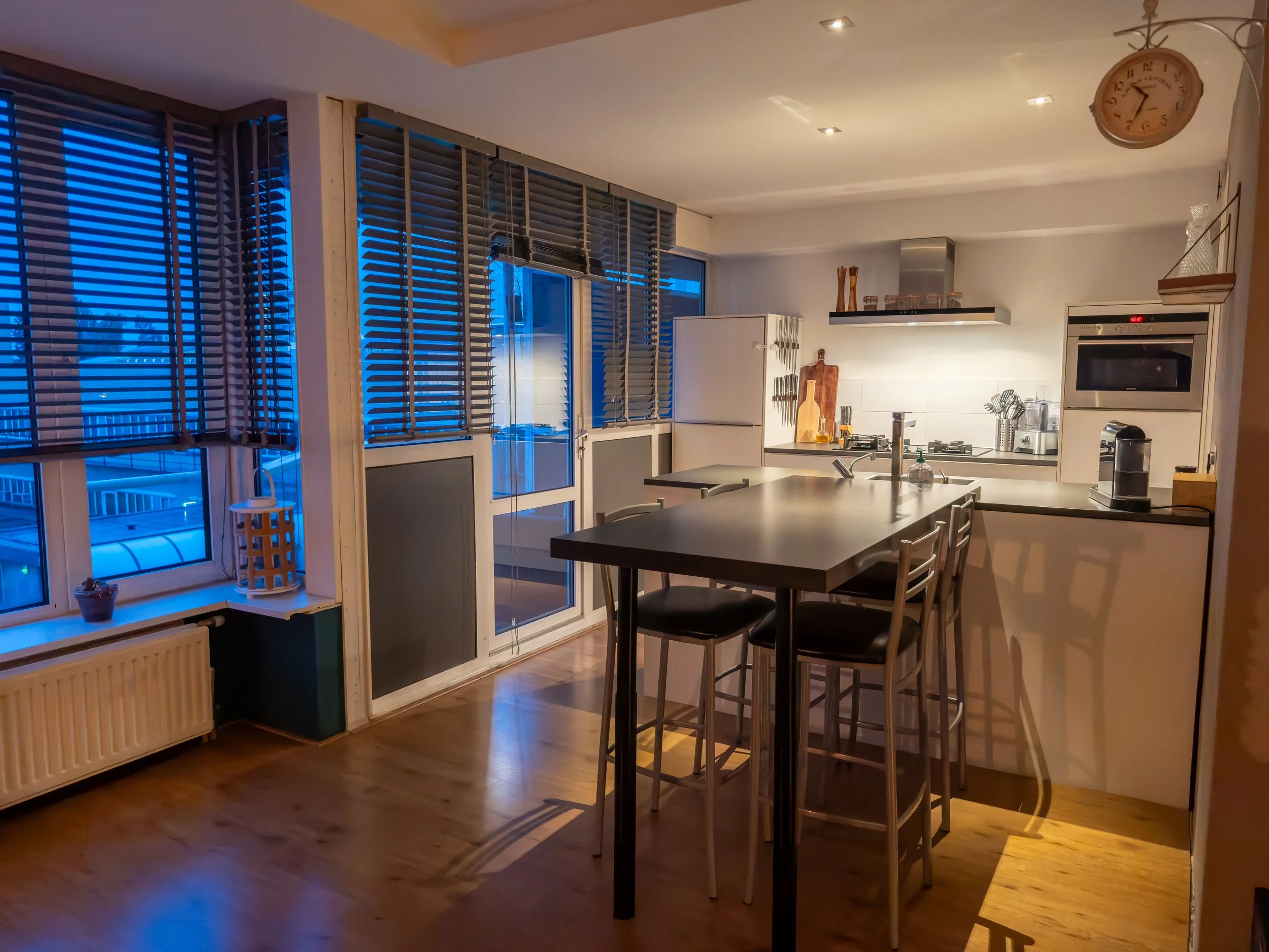 A modern kitchen with a wooden floor, a white refrigerator, a stove on the counter, and a breakfast bar with four black chairs. Large windows with blinds show a blue evening sky. There are various kitchen tools and decor, including a clock hanging on