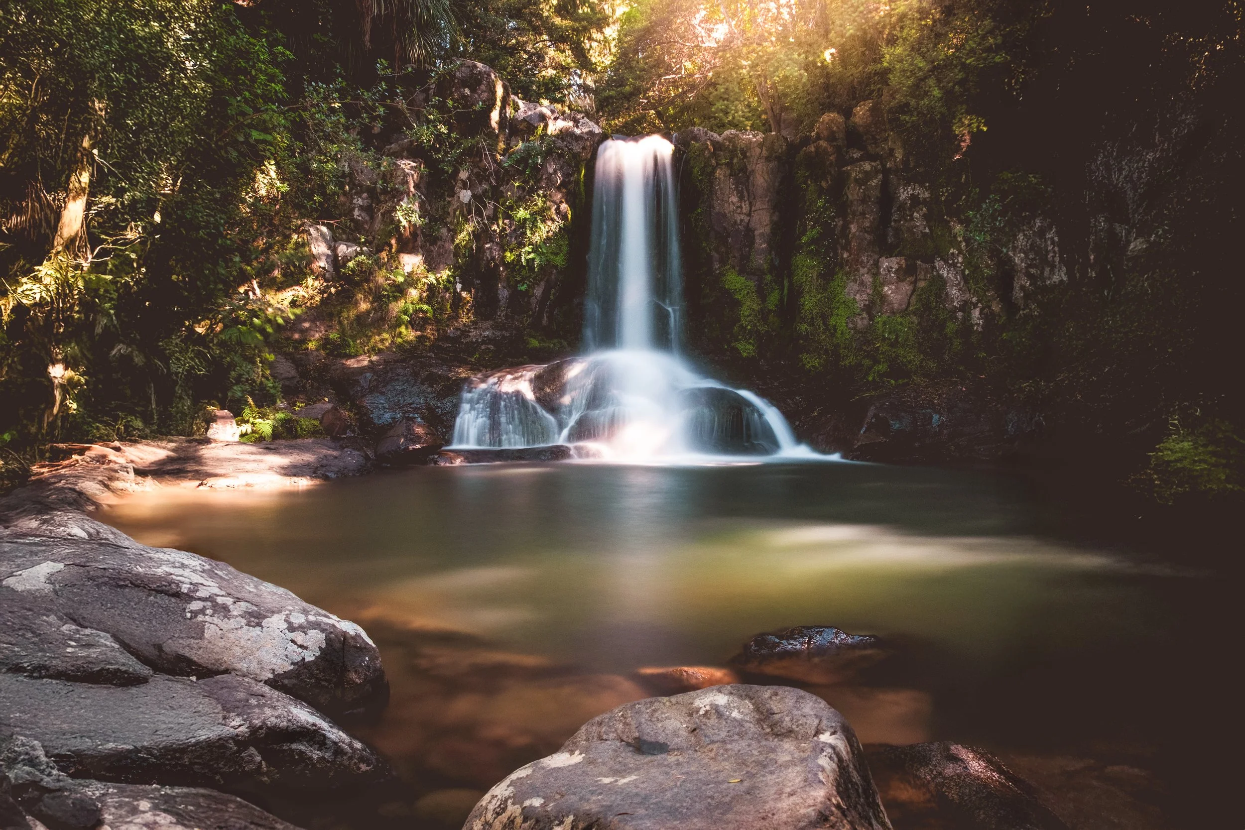 A serene waterfall cascading into a calm pond surrounded by lush green trees and rocks, with sunlight filtering through the foliage.