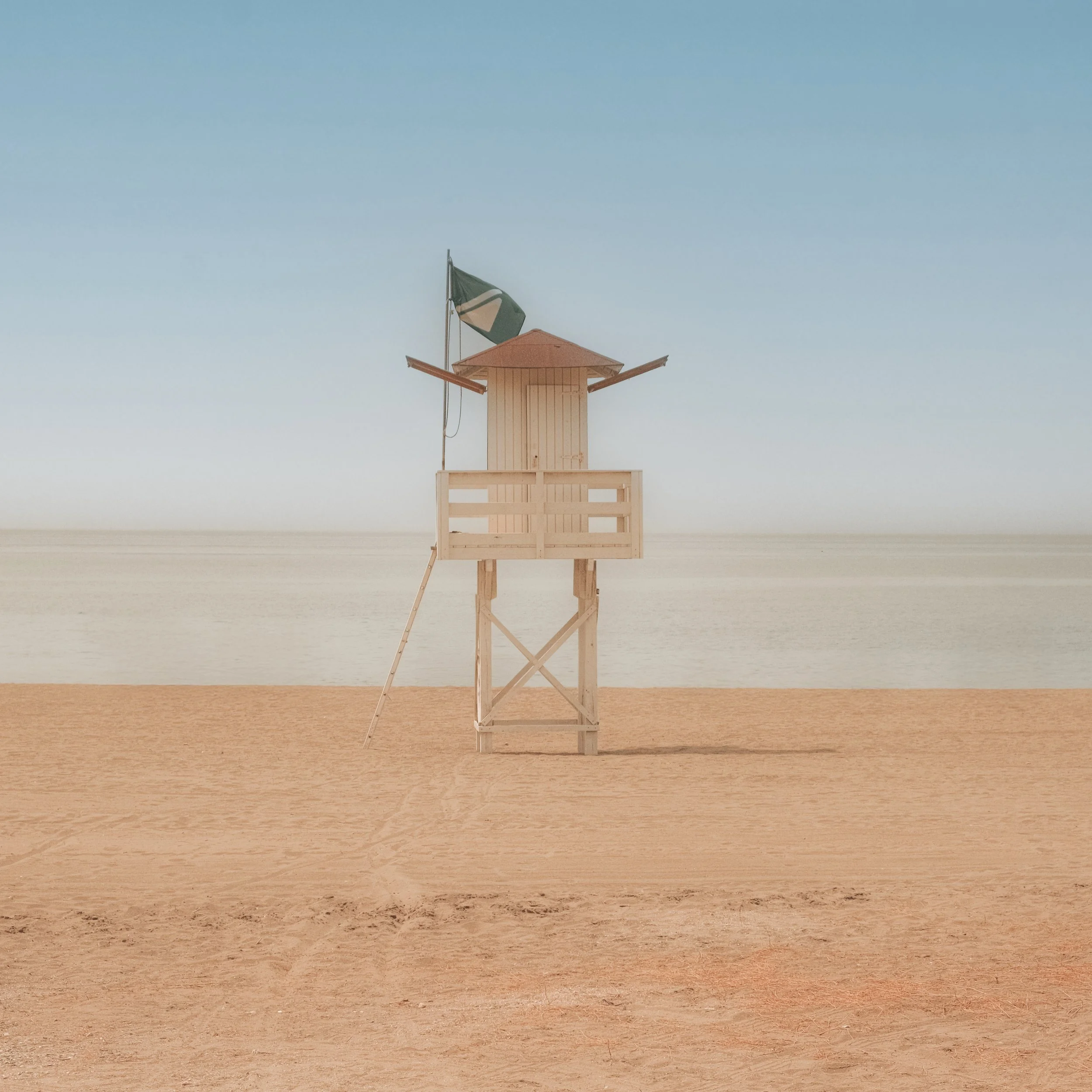 A wooden lifeguard tower on a sandy beach with water and sky in the background. A green and white flag is attached to the tower.