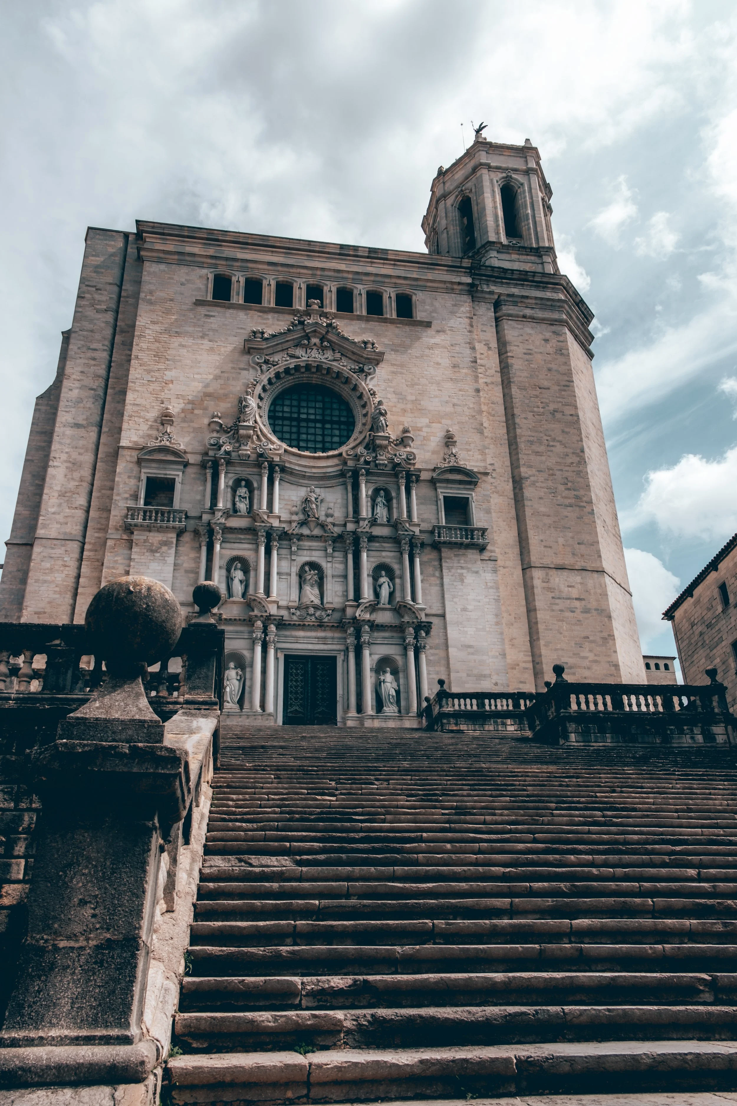 A historic church with baroque architecture, ascending stone steps leading to the entrance, and a cloudy sky in the background.