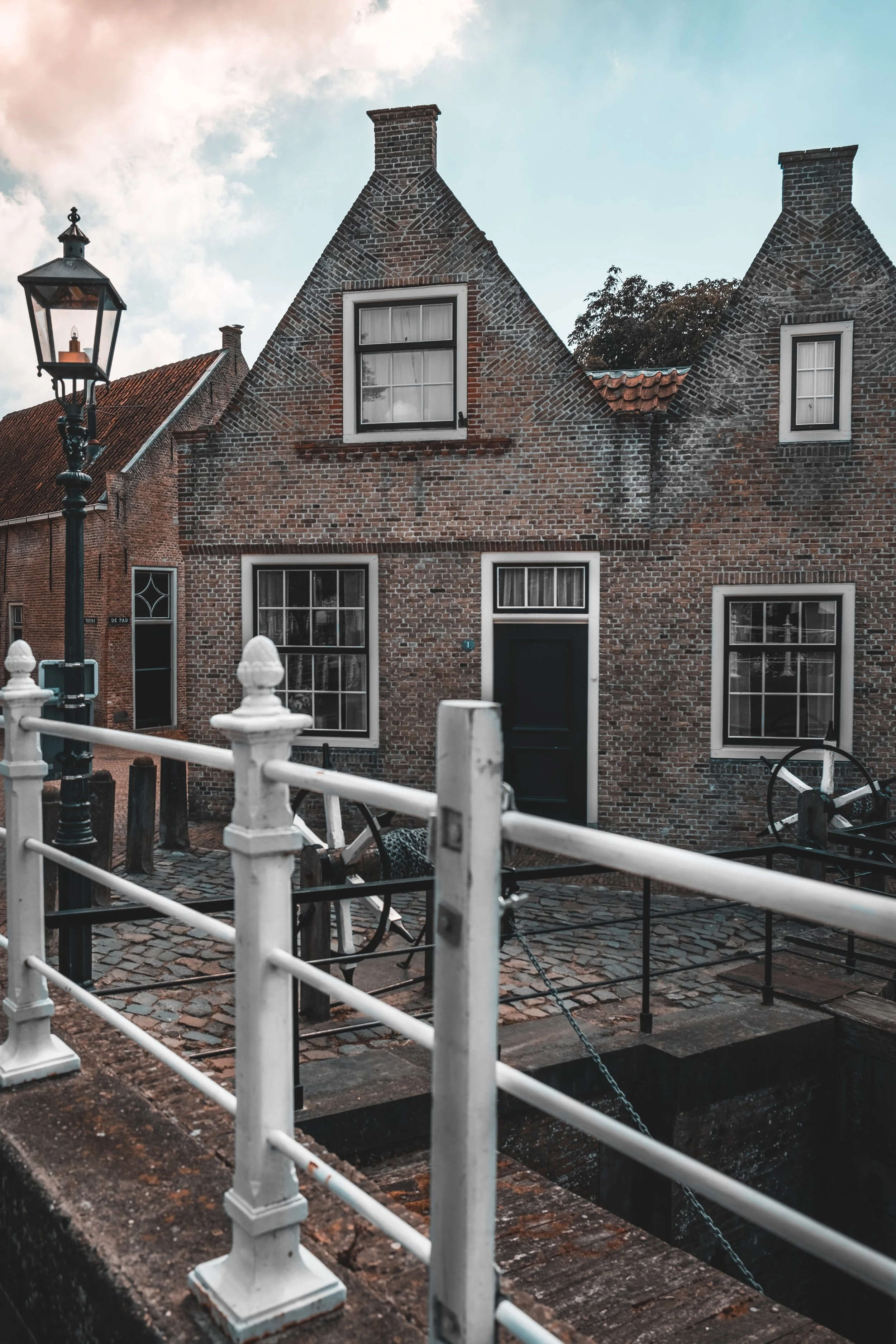 A brick house with white-framed windows and a black door, situated next to a canal with a white fence and an old-fashioned streetlamp, under a partly cloudy sky.