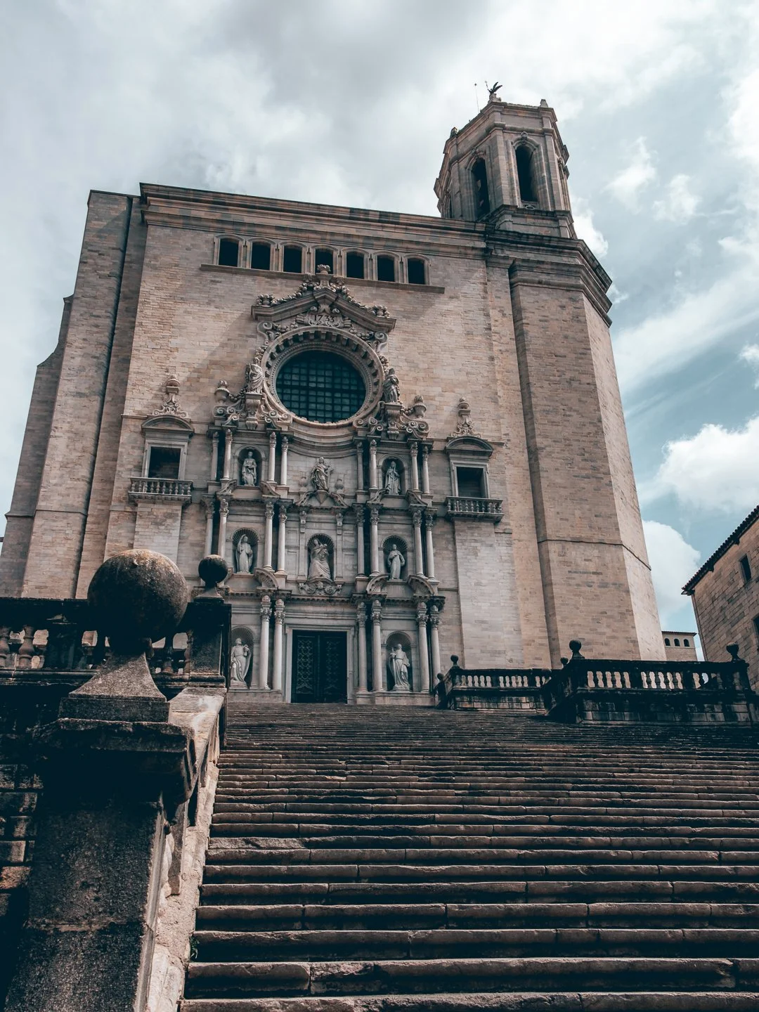 View of a historic church with a grand staircase leading up to the entrance, featuring detailed sculptures and an ornate facade, under a partly cloudy sky. SPAIN GAME OF THRONES GIRONA