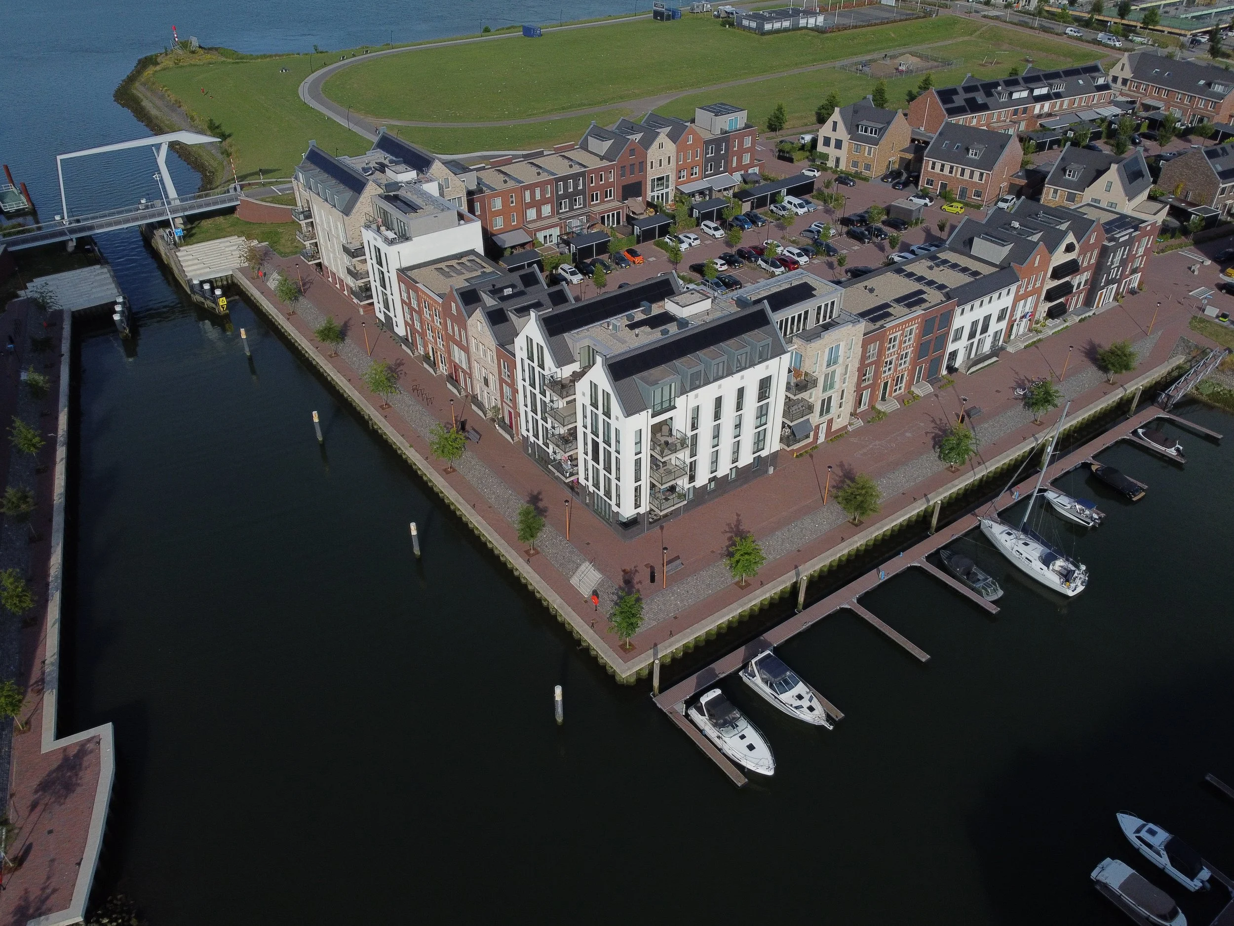 Aerial view of a waterfront residential area with modern apartments, a marina with boats, trees along the sidewalk, and a grassy area with a walking path at the top of the image.